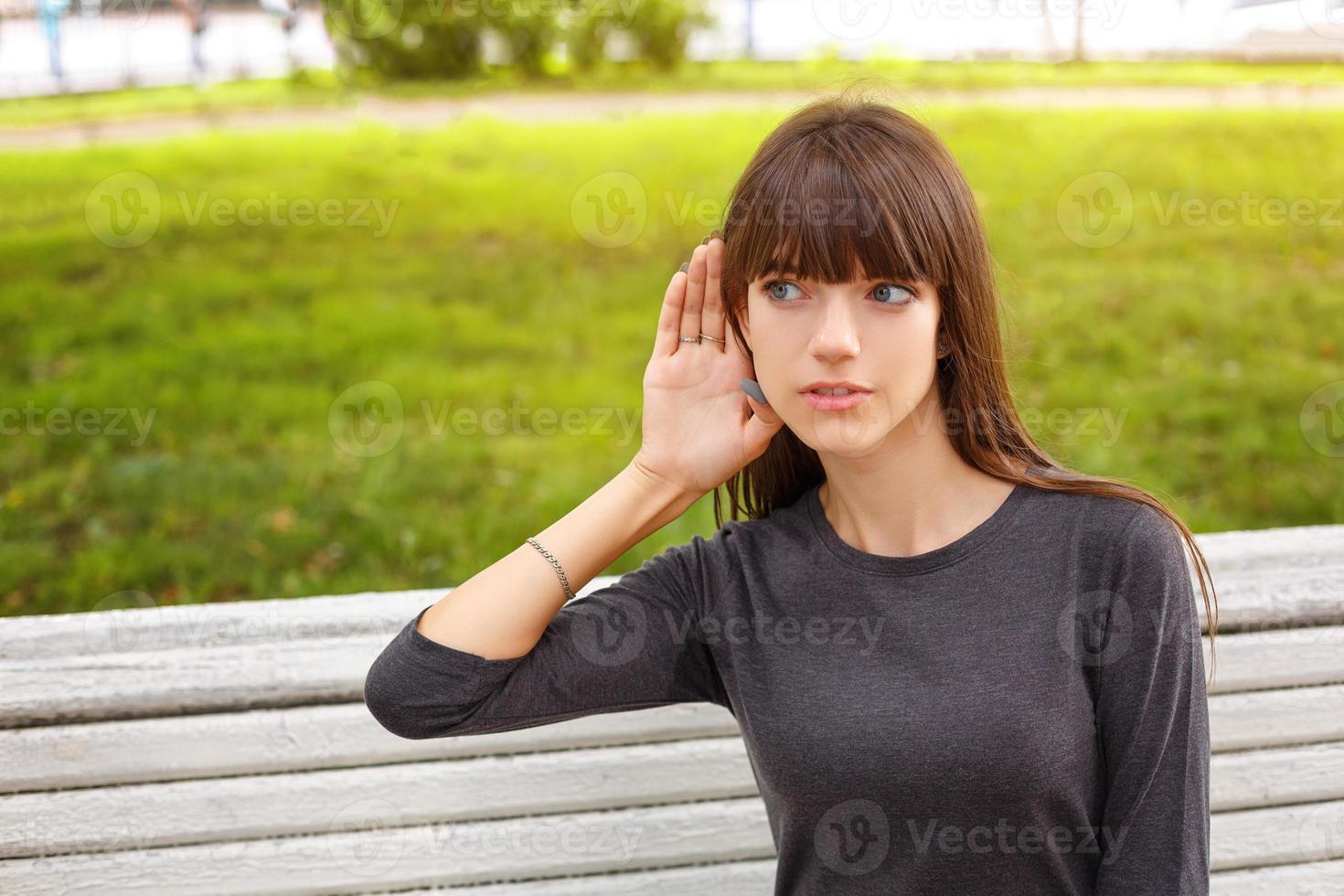 mujer joven en el parque sosteniendo una mano cerca de tu oído, el concepto de escuchar a escondidas foto