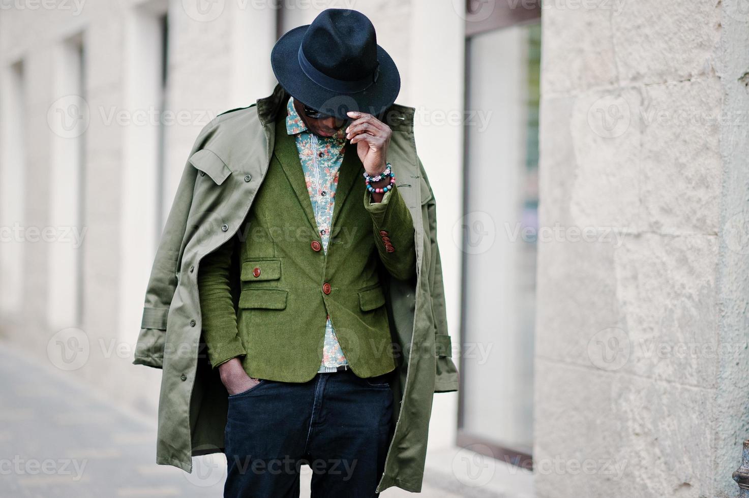 Fashion portrait of black african american man on green velvet jacket, black hat and coat cloak on his shoulders walking on streets of city background house with many windows photo