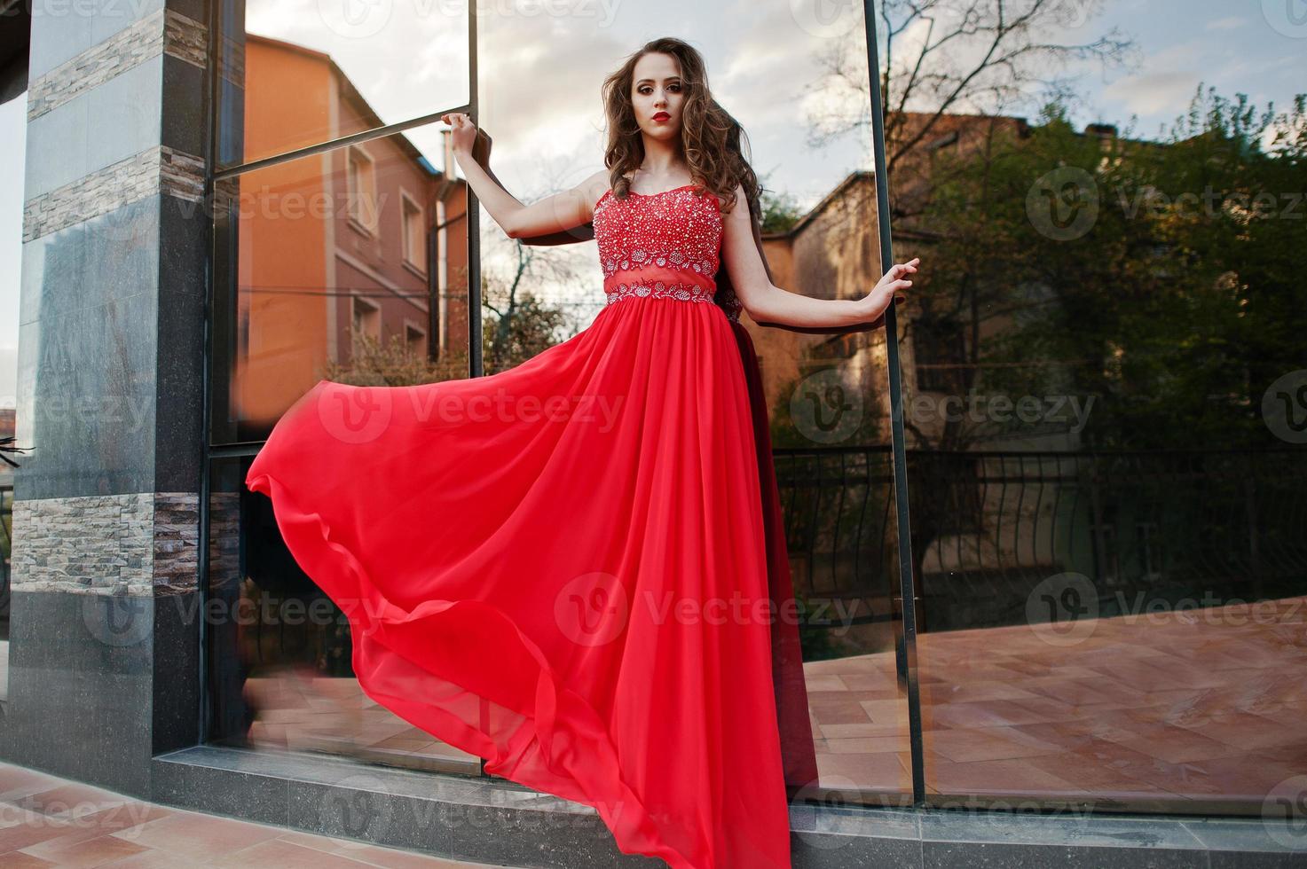 retrato de una chica de moda con vestido de noche rojo posado en la ventana de espejo de fondo de un edificio moderno. vestido que sopla en el aire foto