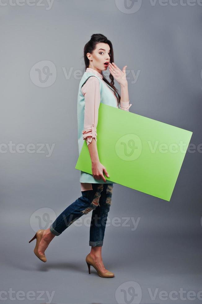 joven con cara sorprendida sostenga papel en blanco verde. joven mujer sonriente mostrar tarjeta en blanco. chica con retrato de pelo largo aislado sobre fondo gris. foto