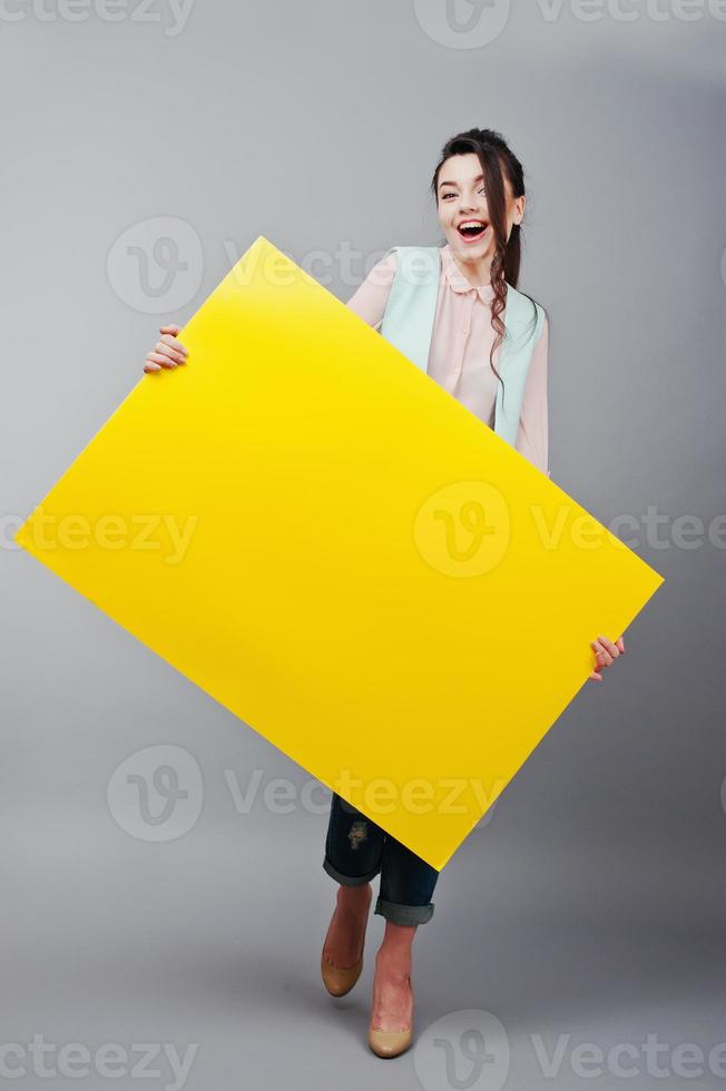 Young girl with surprised face hold yellow blank paper. Young smiling woman show blank card. Girl with long hair portrait isolated on gray background. photo