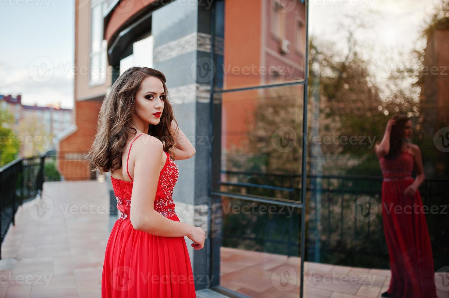 Portrait of fashionable girl at red evening dress posed background mirror window of modern building photo