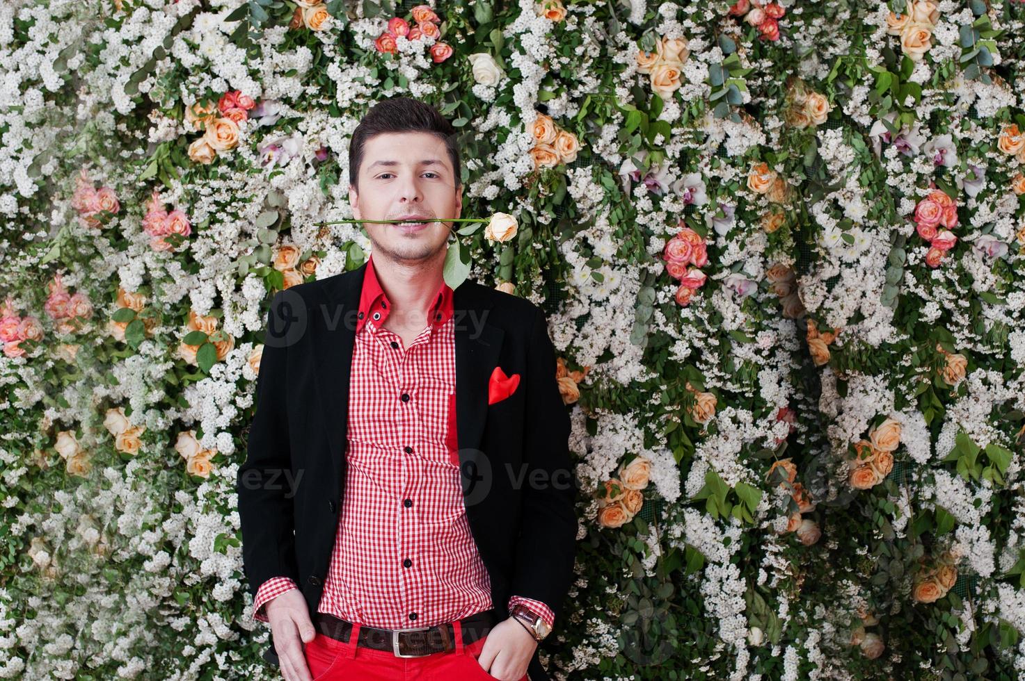 Fashion young man with rose in mouth in black suit and red pants casual poses at studio, background floral decor wall. photo