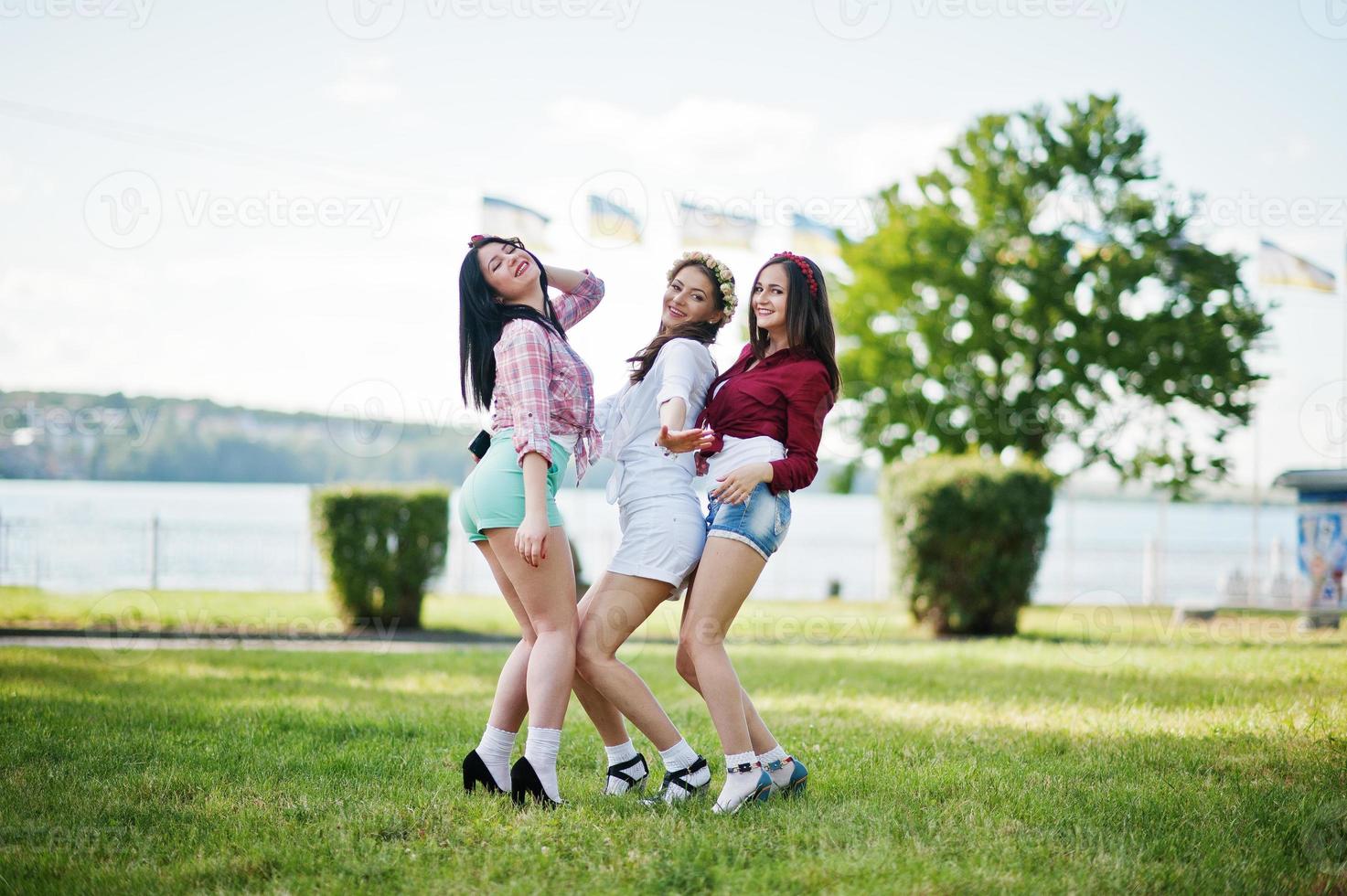 tres niñas felices en pantalones cortos y coronas en la cabeza bailando y divirtiéndose en la hierba verde en la despedida de soltera foto