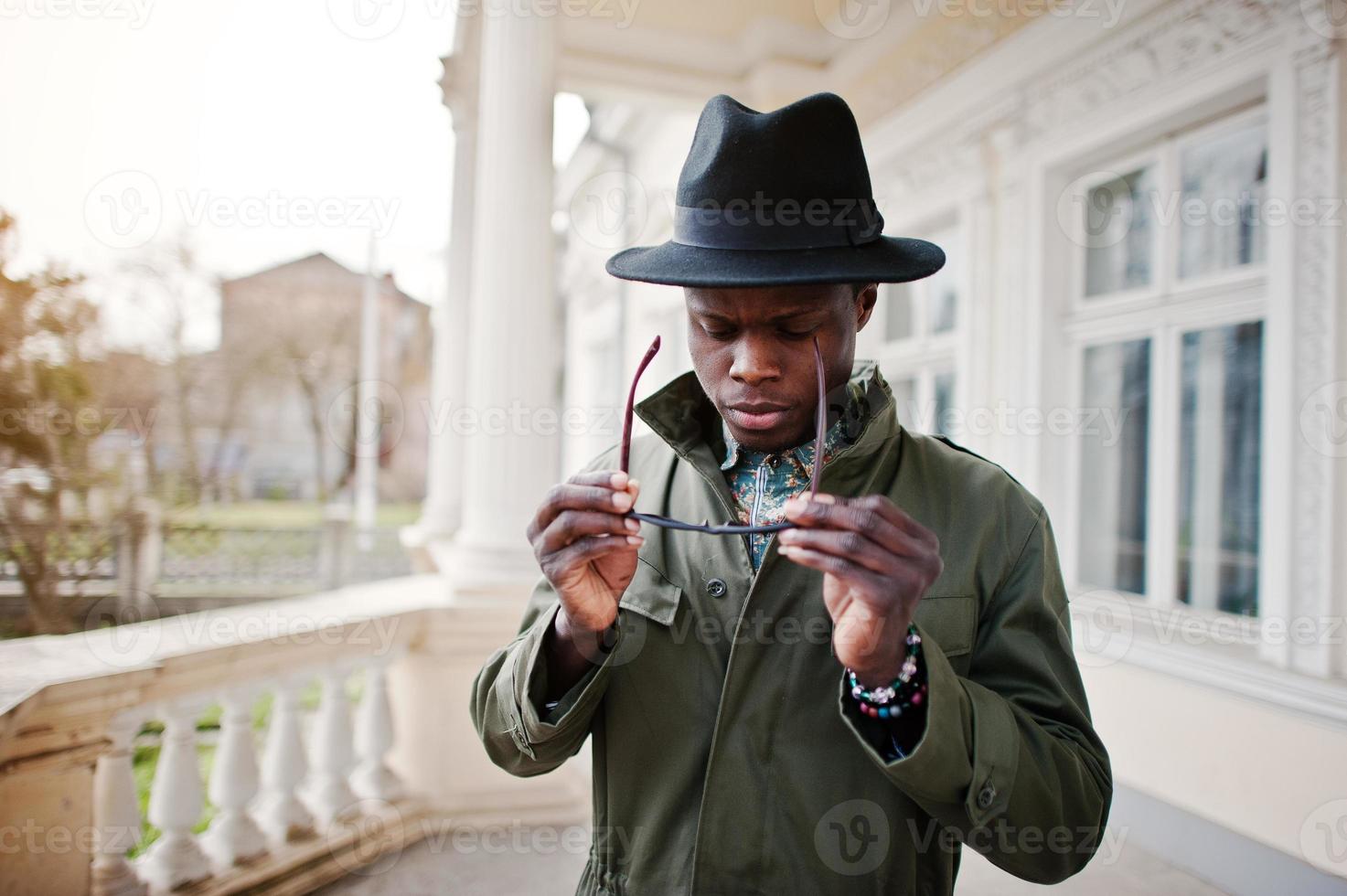 Fashion portrait of black african american man on green coat cloak and black hat, stay on the balcony of yellow mansion photo