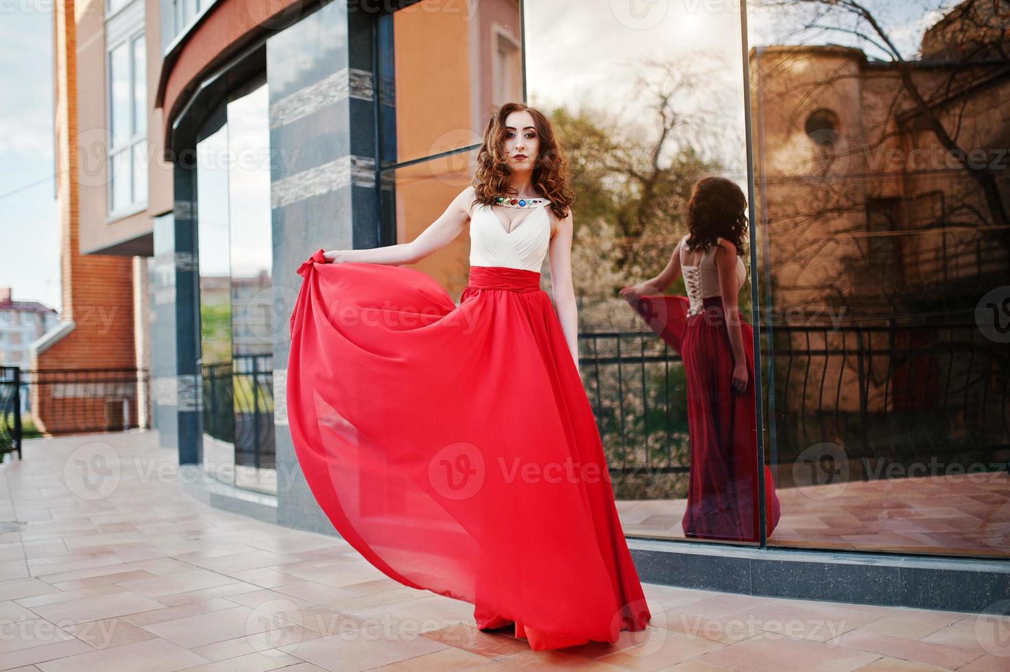 Portrait of fashionable girl at red evening dress posed background mirror window of modern building photo