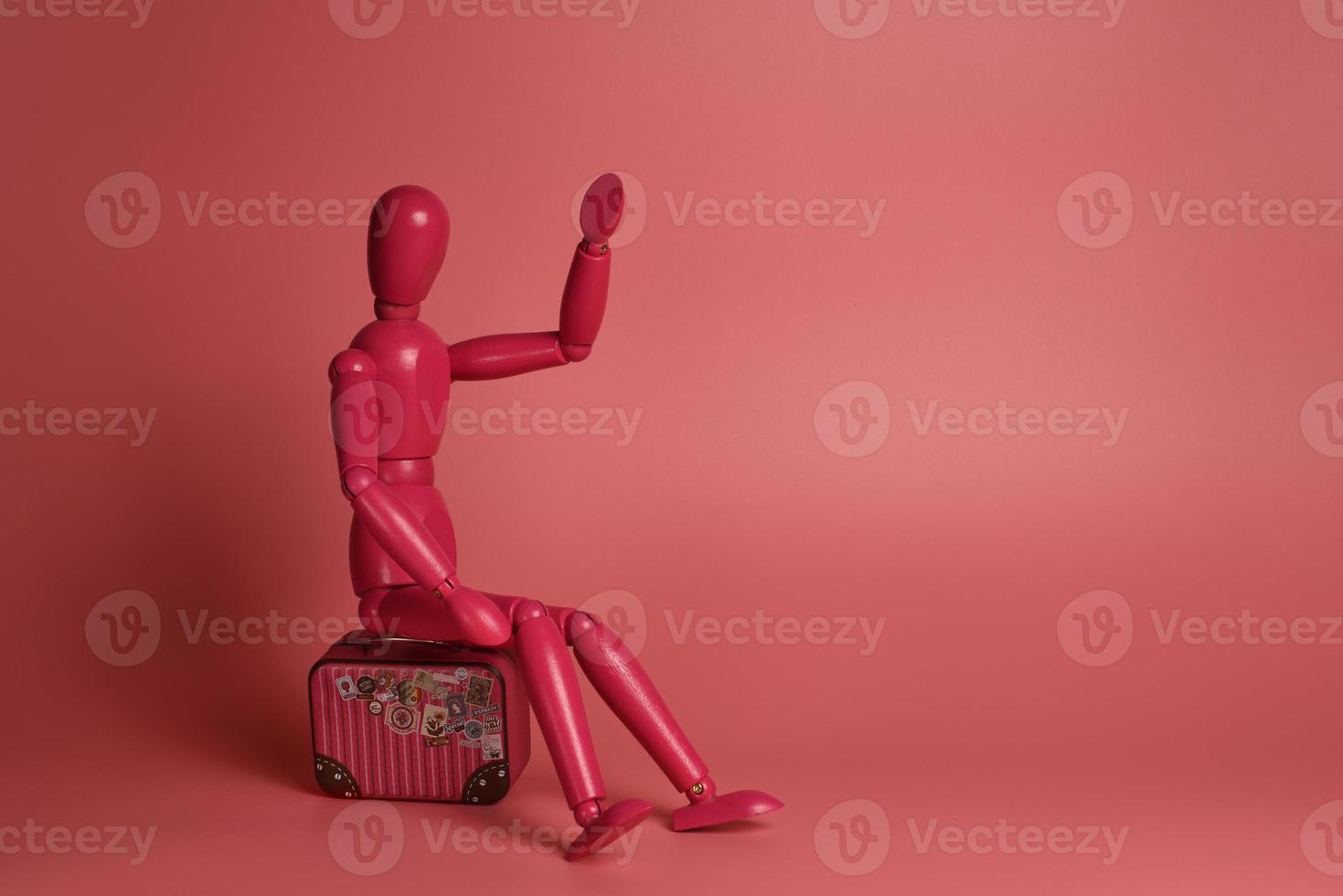 A pink wooden man sits on a suitcase against a pink background. photo