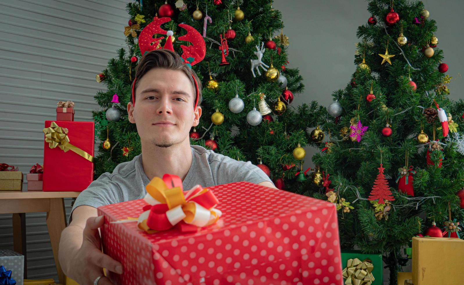 White Caucasian young man, good-looking in concept image, Christmas gift. The young man handed a gift box and looked at the camera. Stack of gift boxes and a Christmas tree decorated in the back. photo