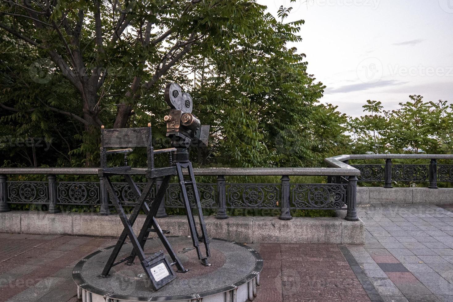 Statue of antique movie camera stands on tripod beside chair with clapboard photo