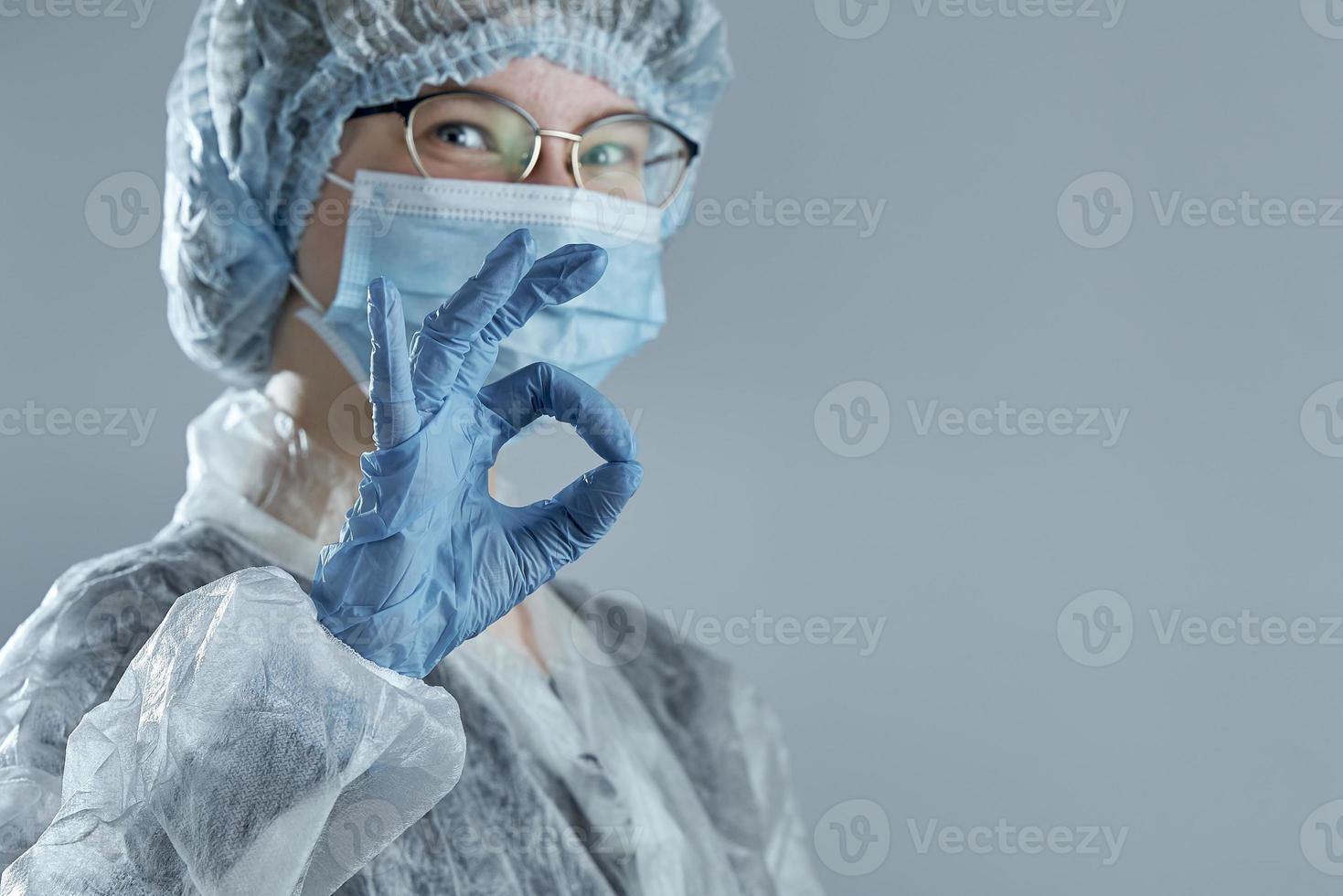 una niña sonriente desdibujada con una máscara médica, guantes de goma y ropa desechable muestra el signo ok. foto