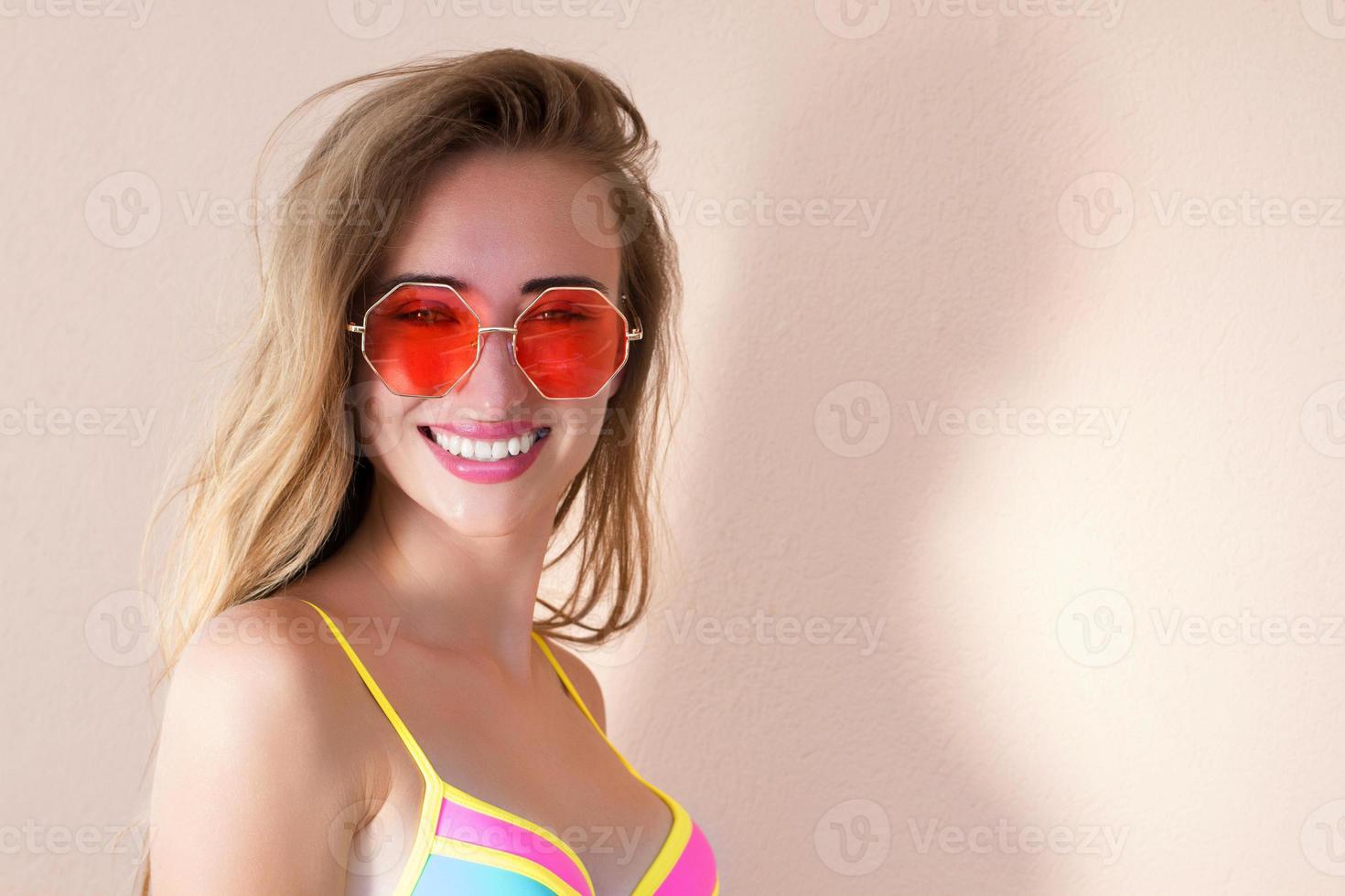 cerca de una chica feliz con gafas de sol rosas aisladas. vacaciones de verano y fin de semana divertido. concepto de verano. mujer joven sonriente en traje de baño de moda. enfoque selectivo. traje de playa de verano. copie el espacio foto