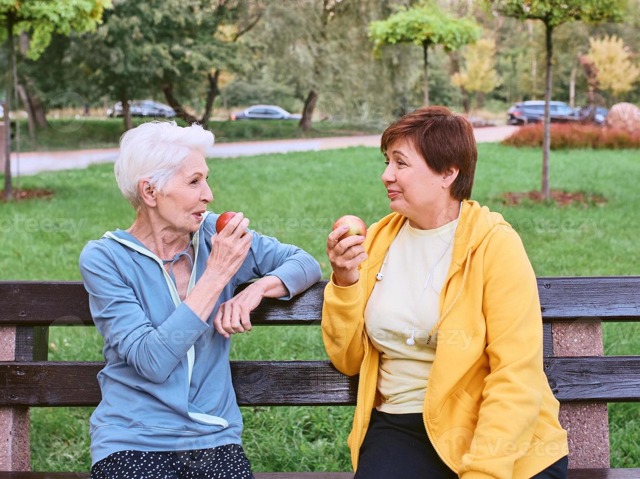 two mature women eating apples on the bench after doing sport exercises in the park. healthy lifestyle concept photo