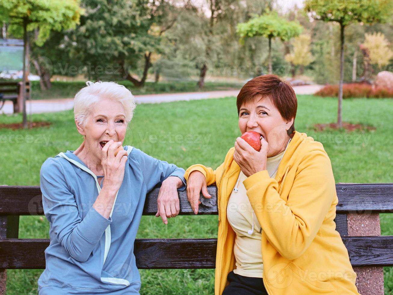 two mature women eating apples on the bench after doing sport exercises in the park. healthy lifestyle concept photo