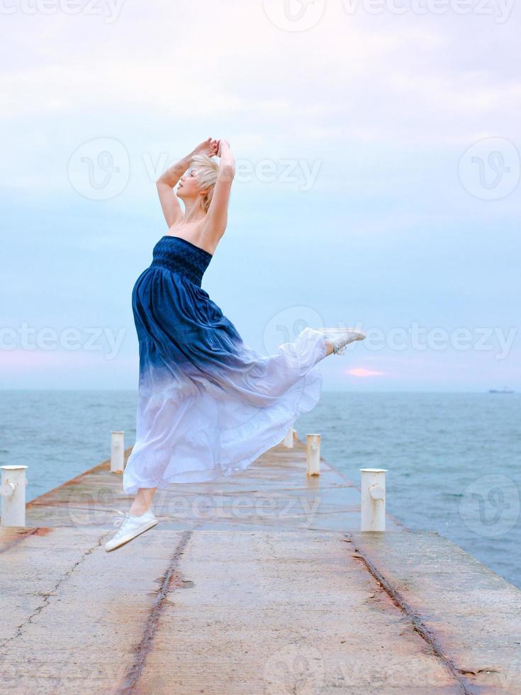 mujer embarazada rubia blanca caucásica con pelo corto en vestido largo y zapatillas felices de estar embarazada y saltando junto al mar al amanecer foto