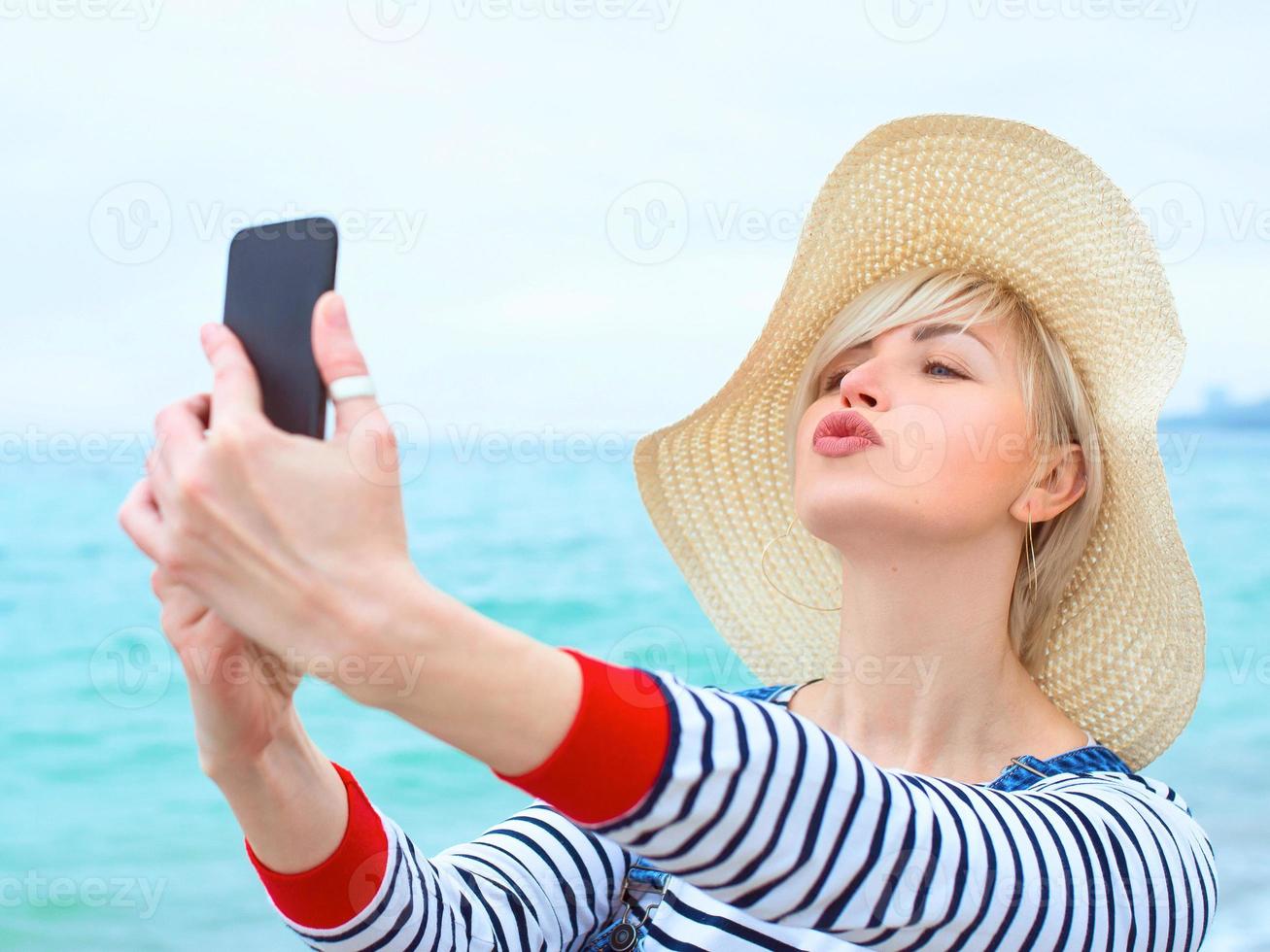 hermosa joven rubia caucásica de vacaciones con sombrero de paja, blusa a rayas y overoles de mezclilla haciendo selfie en el teléfono inteligente junto al increíble fondo azul del mar foto