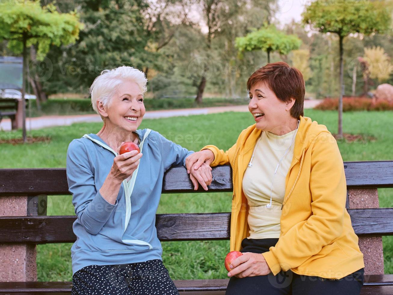 two mature women eating apples on the bench after doing sport exercises in the park. healthy lifestyle concept photo