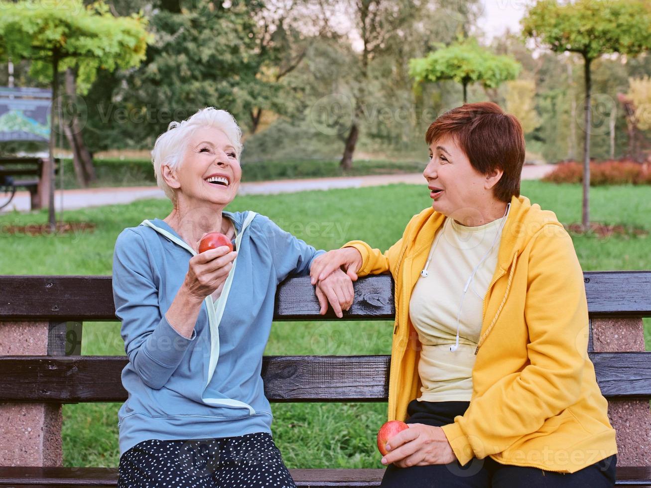 dos mujeres maduras comiendo manzanas en el banco después de hacer ejercicios deportivos en el parque. concepto de estilo de vida saludable foto