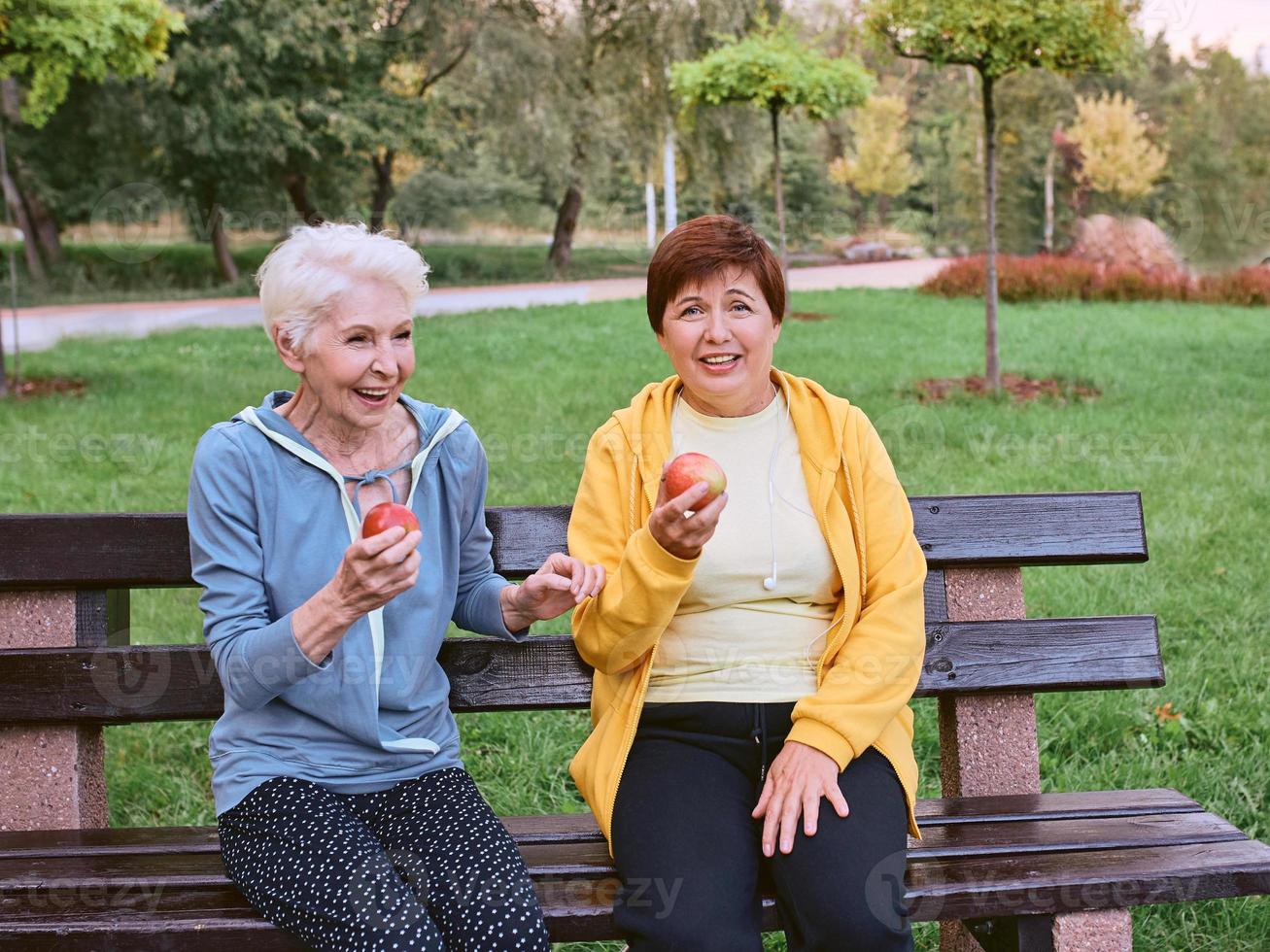 dos mujeres maduras comiendo manzanas en el banco después de hacer ejercicios deportivos en el parque. concepto de estilo de vida saludable foto