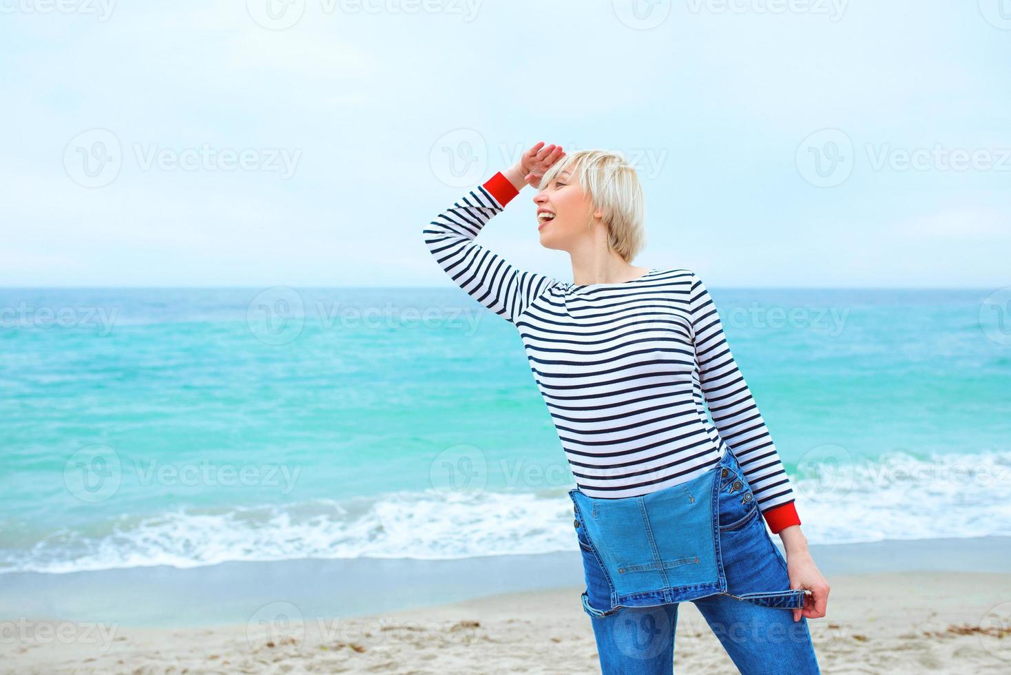 beautiful young blonde caucasian woman on vacation in striped blouse, sneakers and denim overall on the beach by the amazing blue sea background. Happy to be at the sea photo