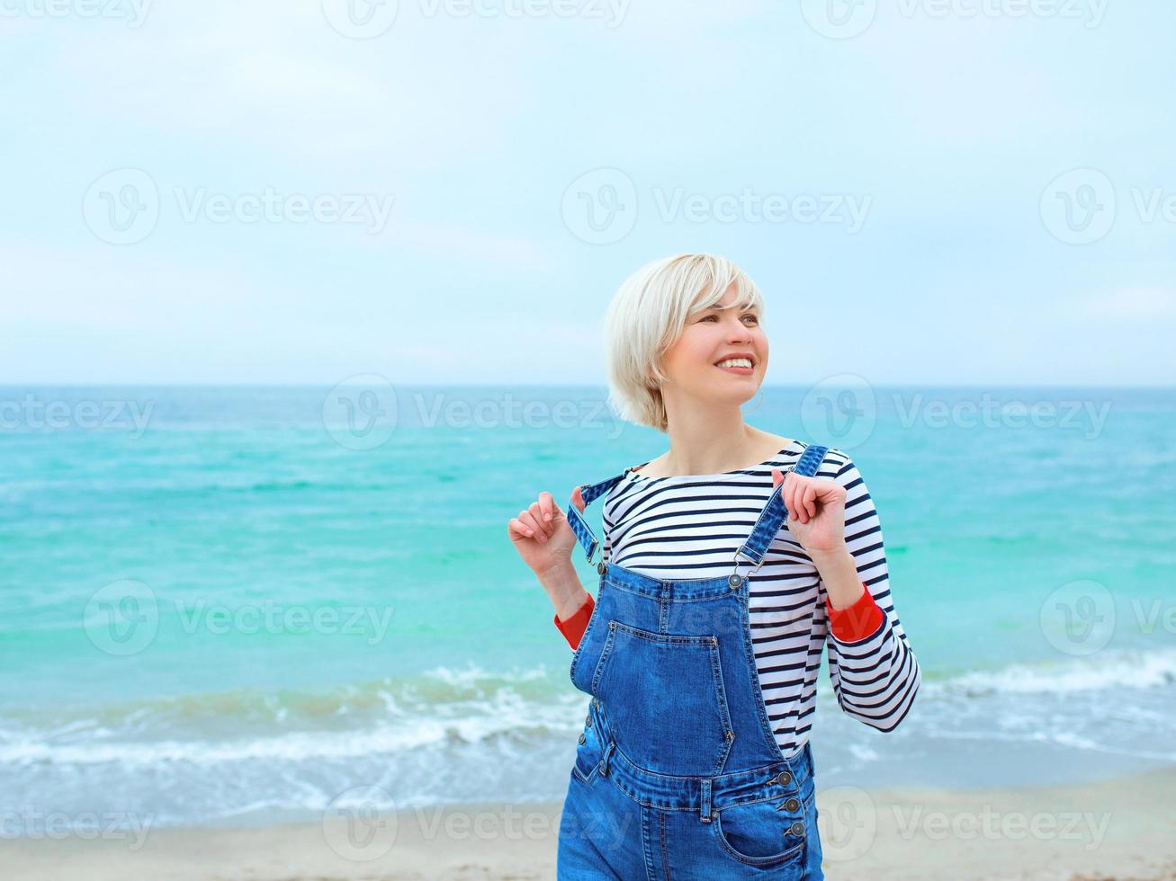 beautiful young blonde caucasian woman on vacation in striped blouse, sneakers and denim overall on the beach by the amazing blue sea background. Happy to be at the sea photo