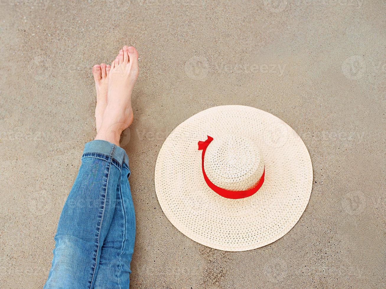 legs and feet of caucasian woman in jeans sitting by the sea on the coast with hat near by on sand background. Travel abstract photo