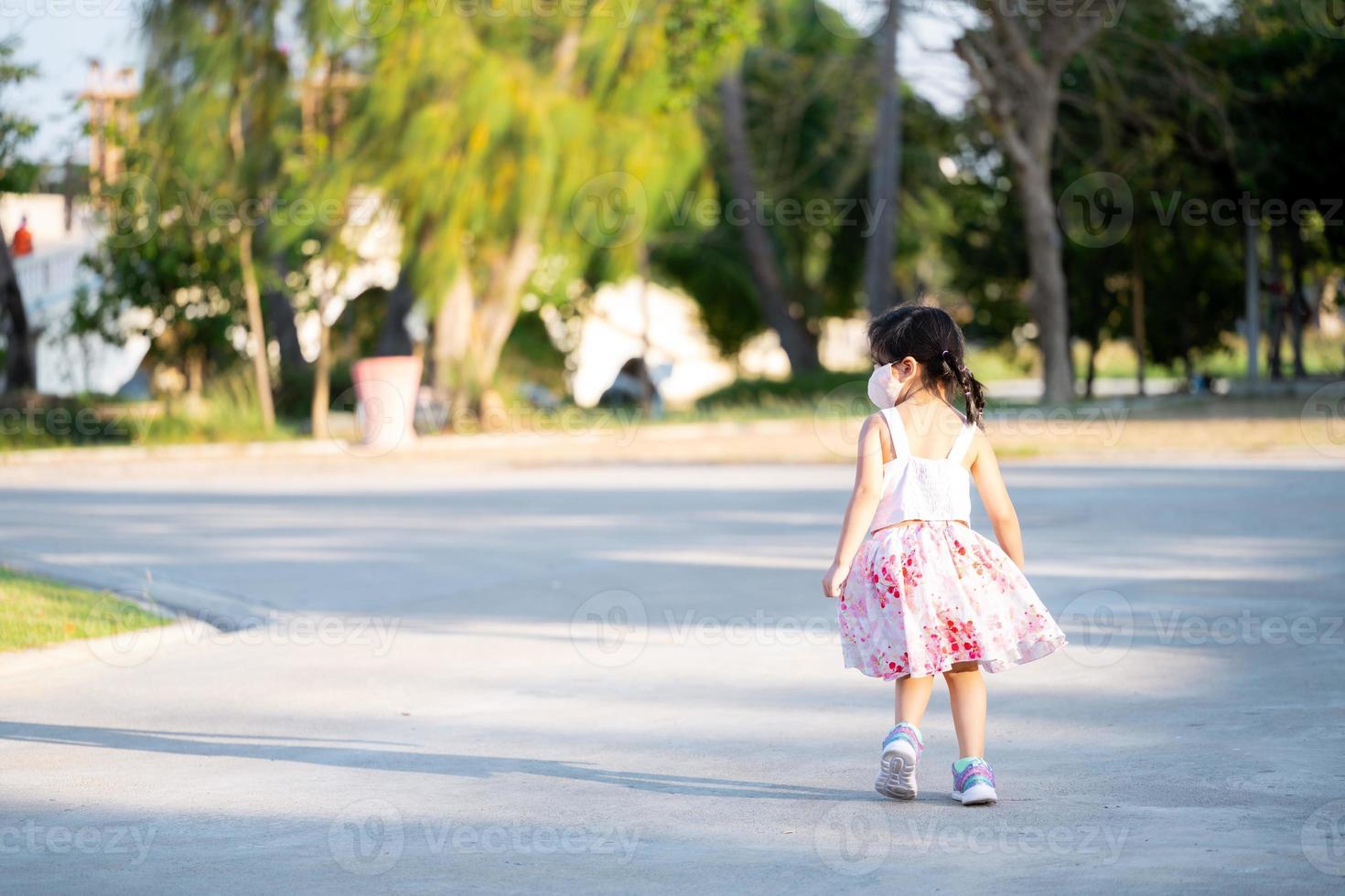 Rear back view of kid girl is jogging in evening at public park. Child wear cloth face mask that can insert filters for PM2.5 dust and prevent viruses. photo