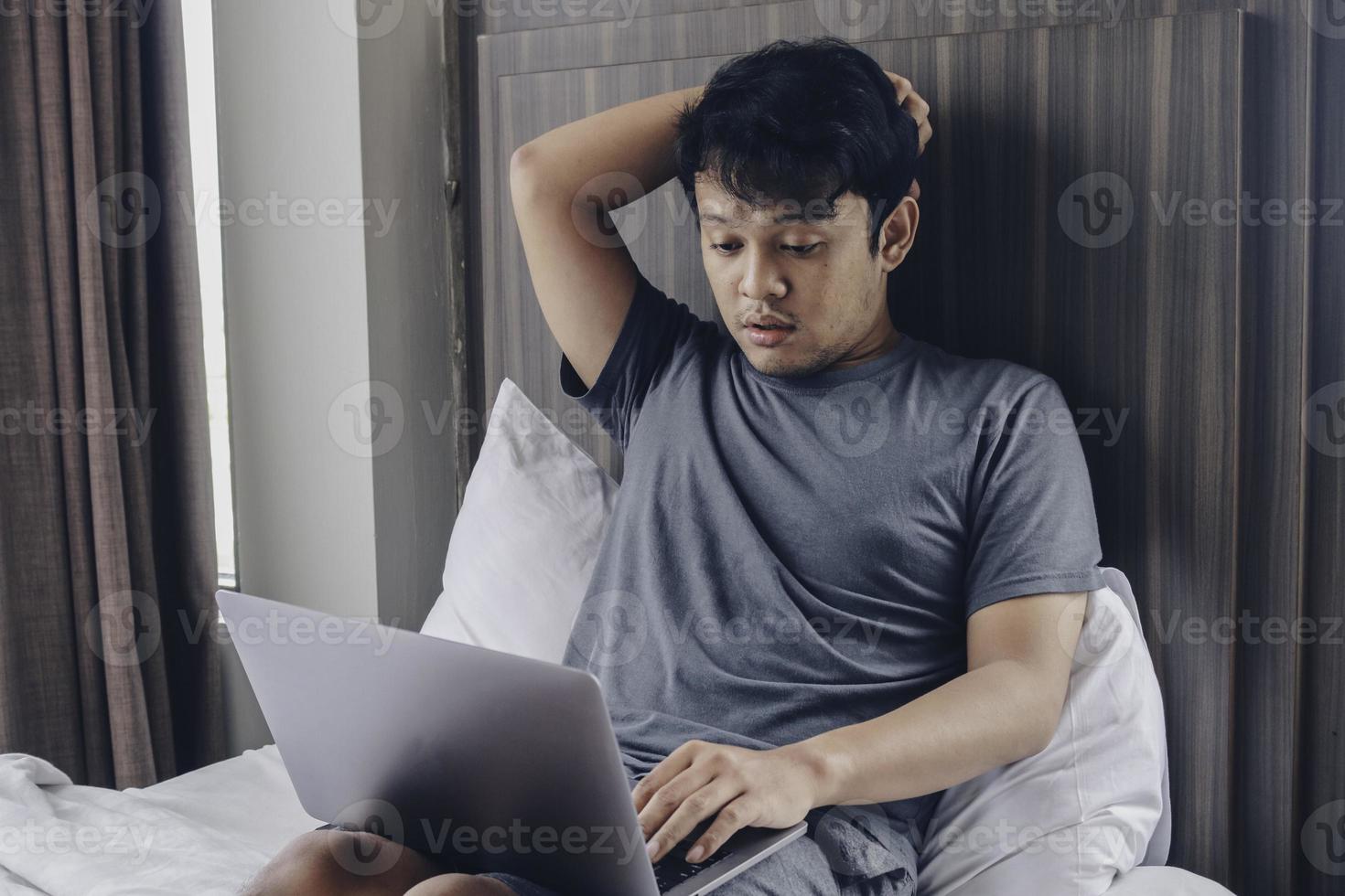 Tired stressed young Asian man feeling sleepy and tired while using laptop on the bed in bedroom. Hard work concept. photo