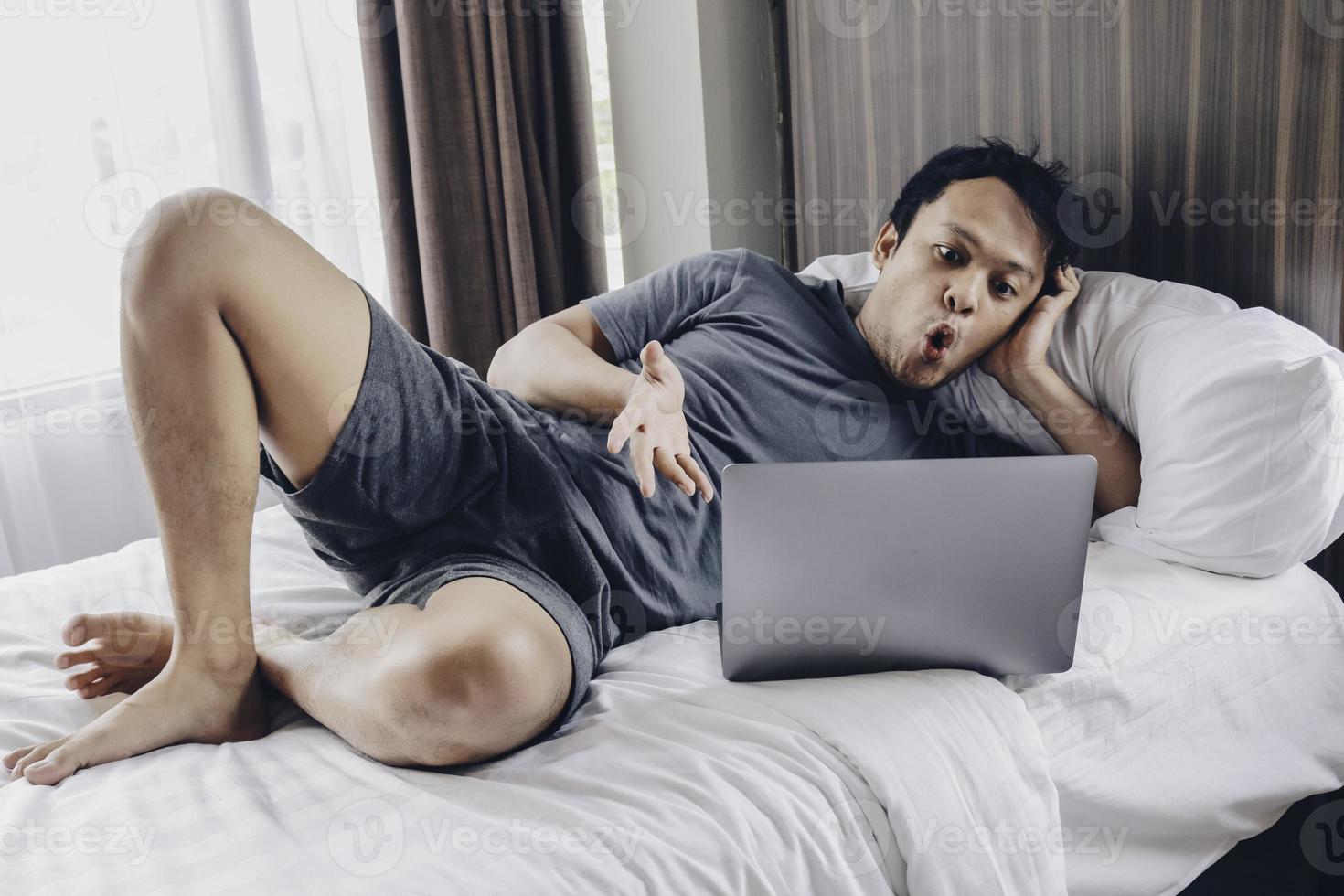 un hombre asiático sorprendido y conmocionado está trabajando con su computadora portátil en su acogedora cama. concepto de estilo de vida freelance exitoso. foto