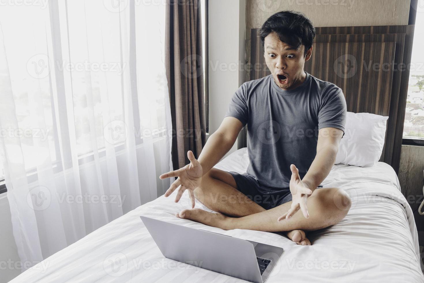 un hombre asiático sorprendido y conmocionado está trabajando con su computadora portátil en su acogedora cama. concepto de estilo de vida freelance exitoso. foto