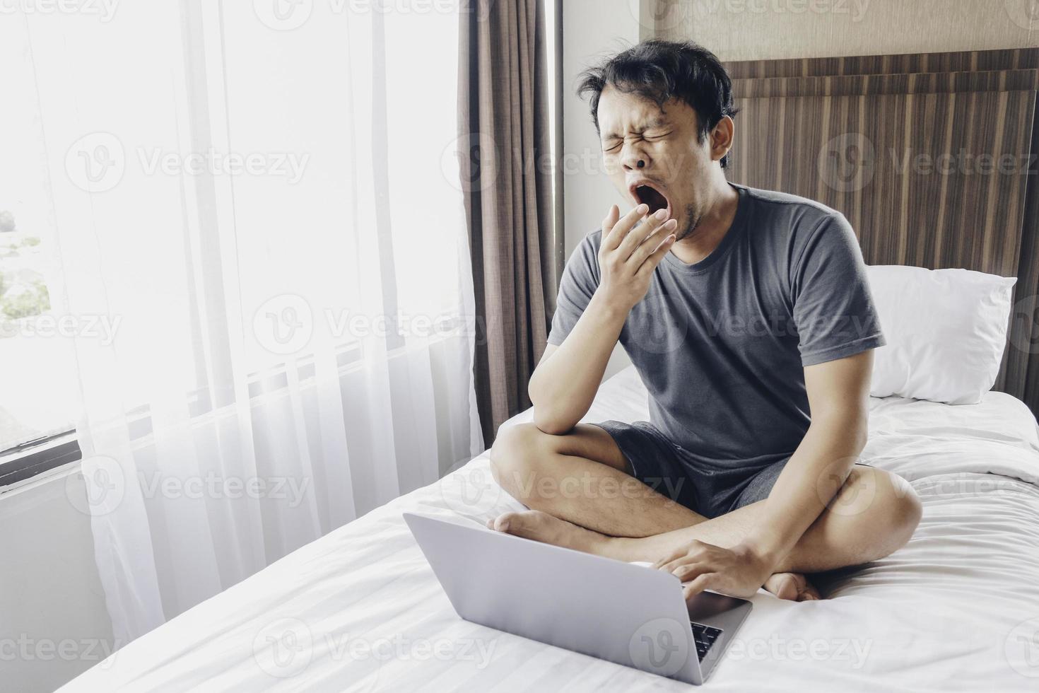 Tired stressed young Asian man feeling sleepy and tired while using laptop on the bed in bedroom. Hard work concept. photo