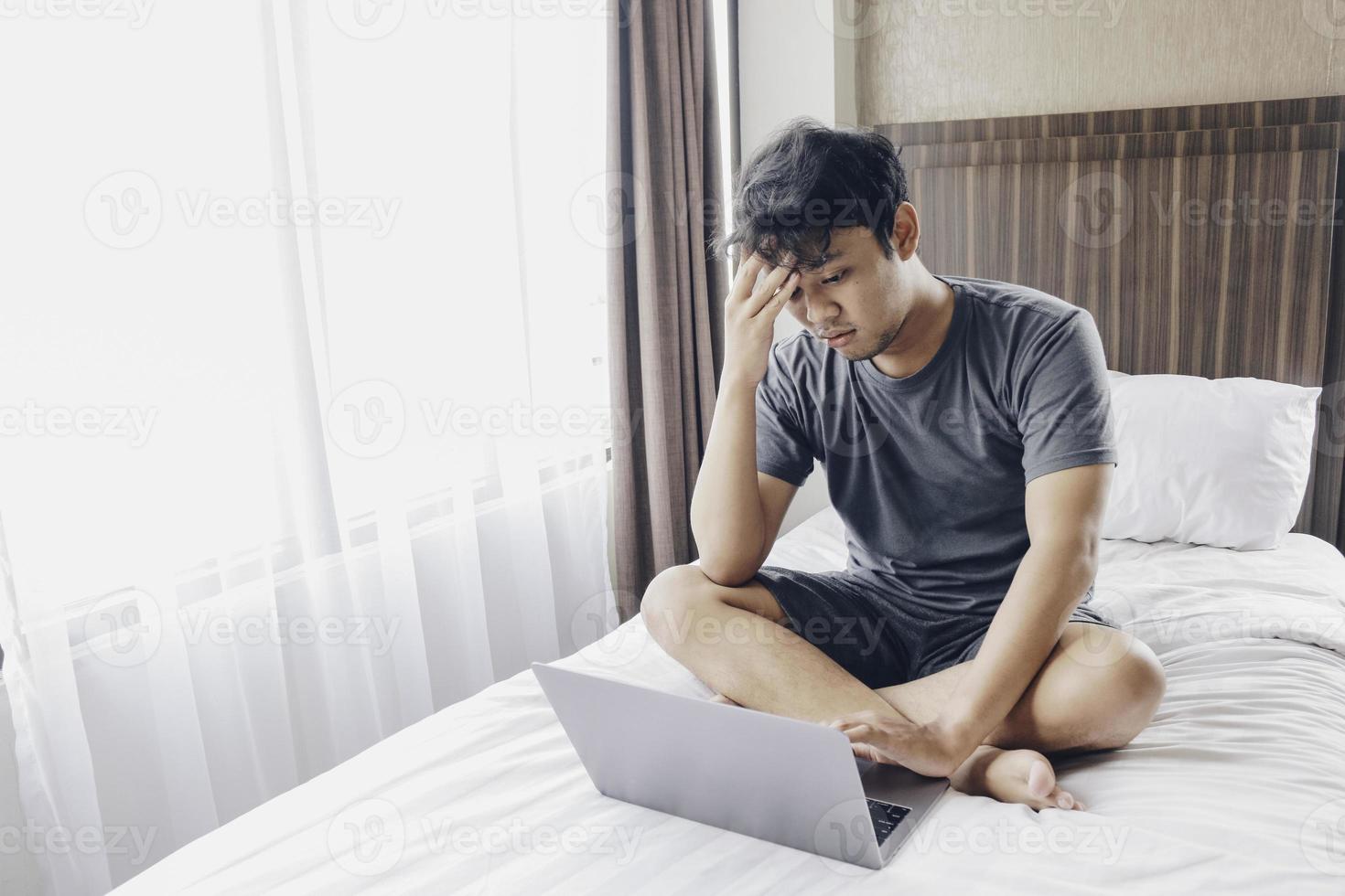 Tired stressed young Asian man feeling sleepy and tired while using laptop on the bed in bedroom. Hard work concept. photo