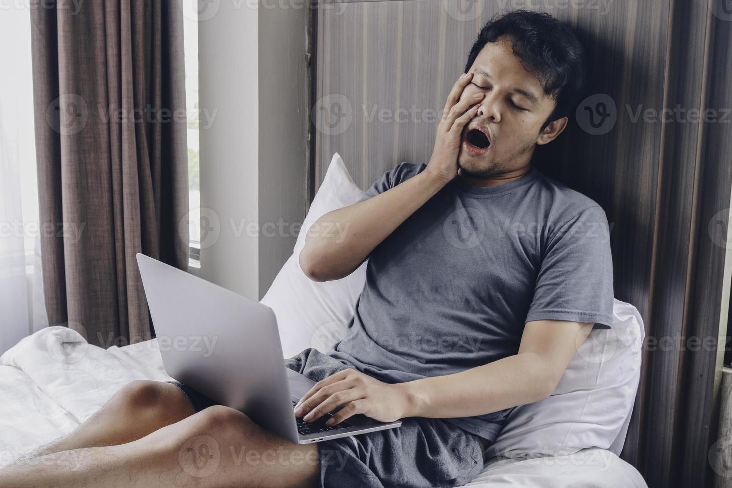 Tired stressed young Asian man feeling sleepy and tired while using laptop on the bed in bedroom. Hard work concept. photo