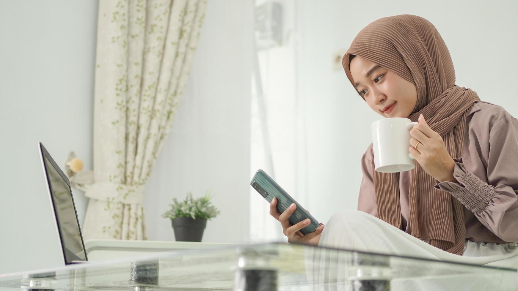 beautiful woman in hijab working from home looking for good ideas on her smartphone while enjoying a drink photo