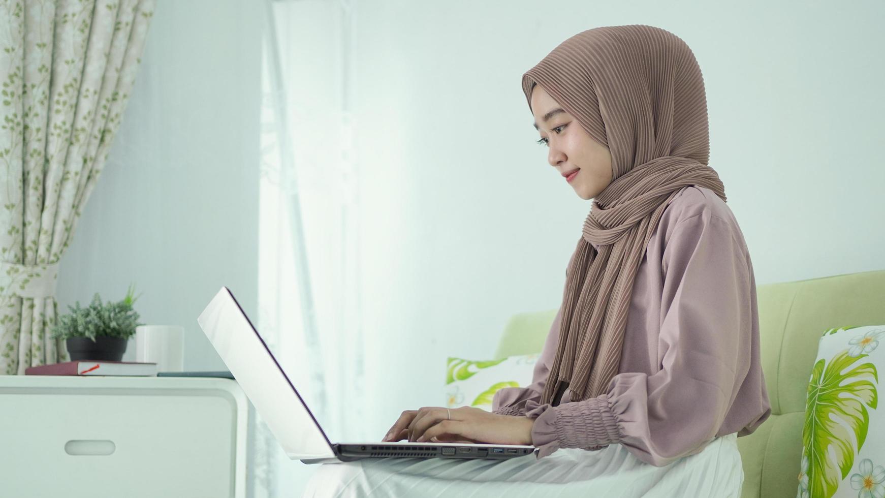asian woman in hijab sitting enjoying doing something on her laptop photo