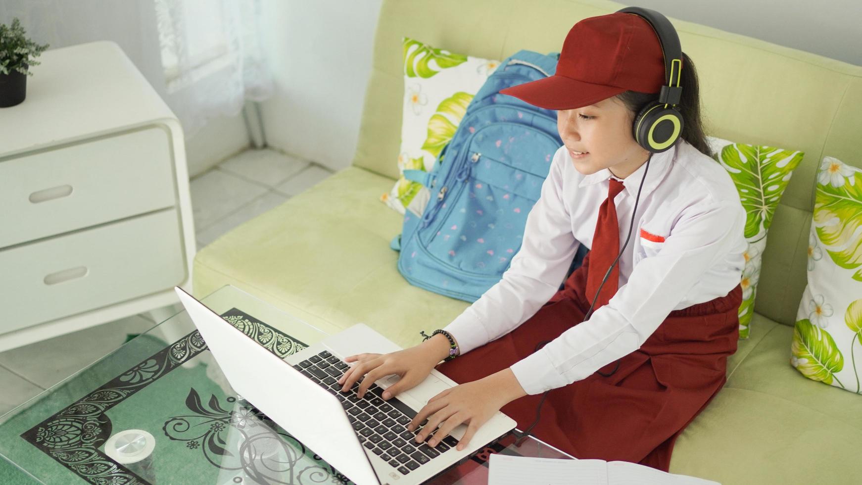 niña asiática de la escuela primaria que estudia en línea en casa escribiendo mientras escucha foto