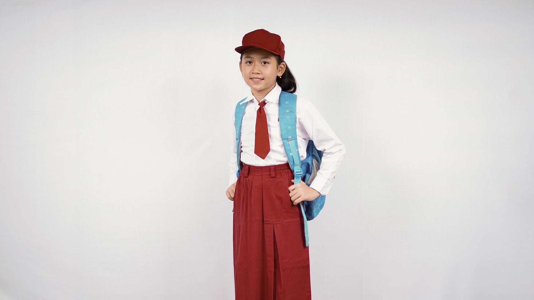 niña asiática con sombrero y mochila escolar sonriendo alegremente con fondo blanco aislado foto