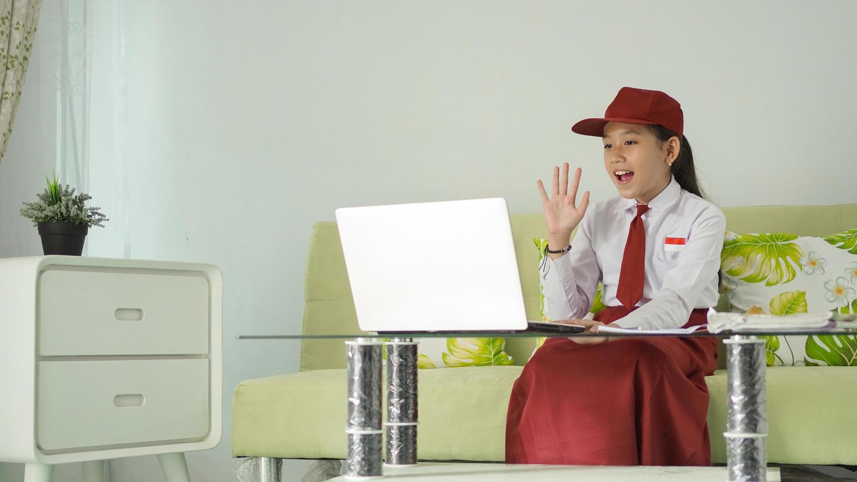 asian elementary school girl studying from home saying hello to her laptop screen photo