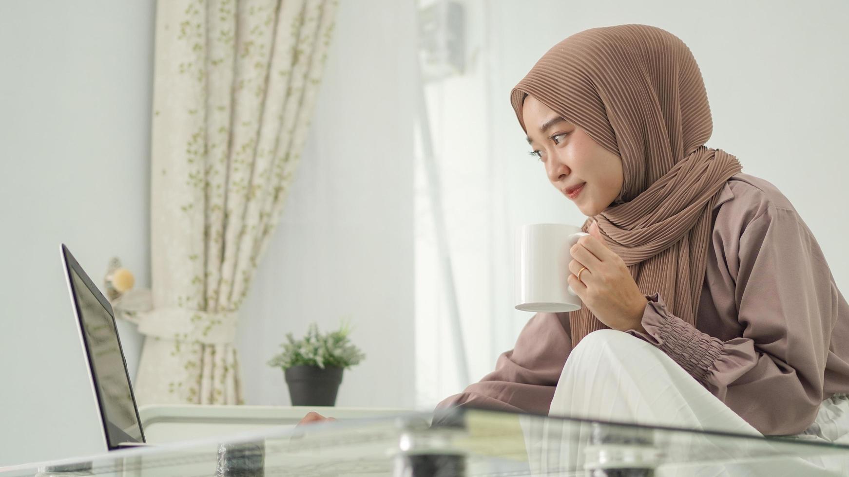 beautiful woman in hijab working from home using laptop while enjoying a drink photo