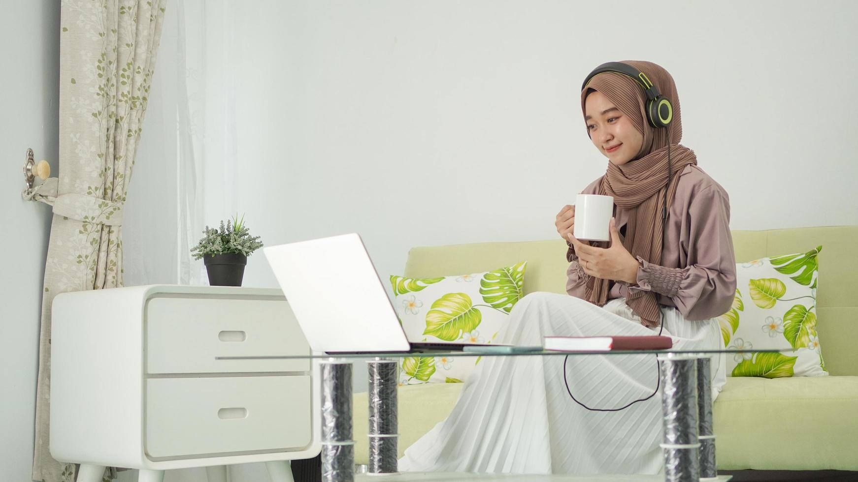 young woman in hijab working from home enjoying a drink while listening on headphones photo