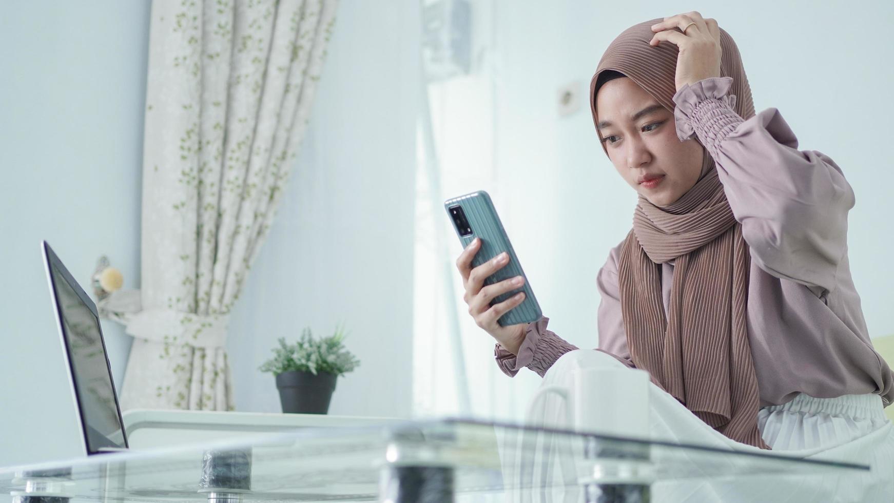 asian woman in hijab looking at phone screen frantically at home photo