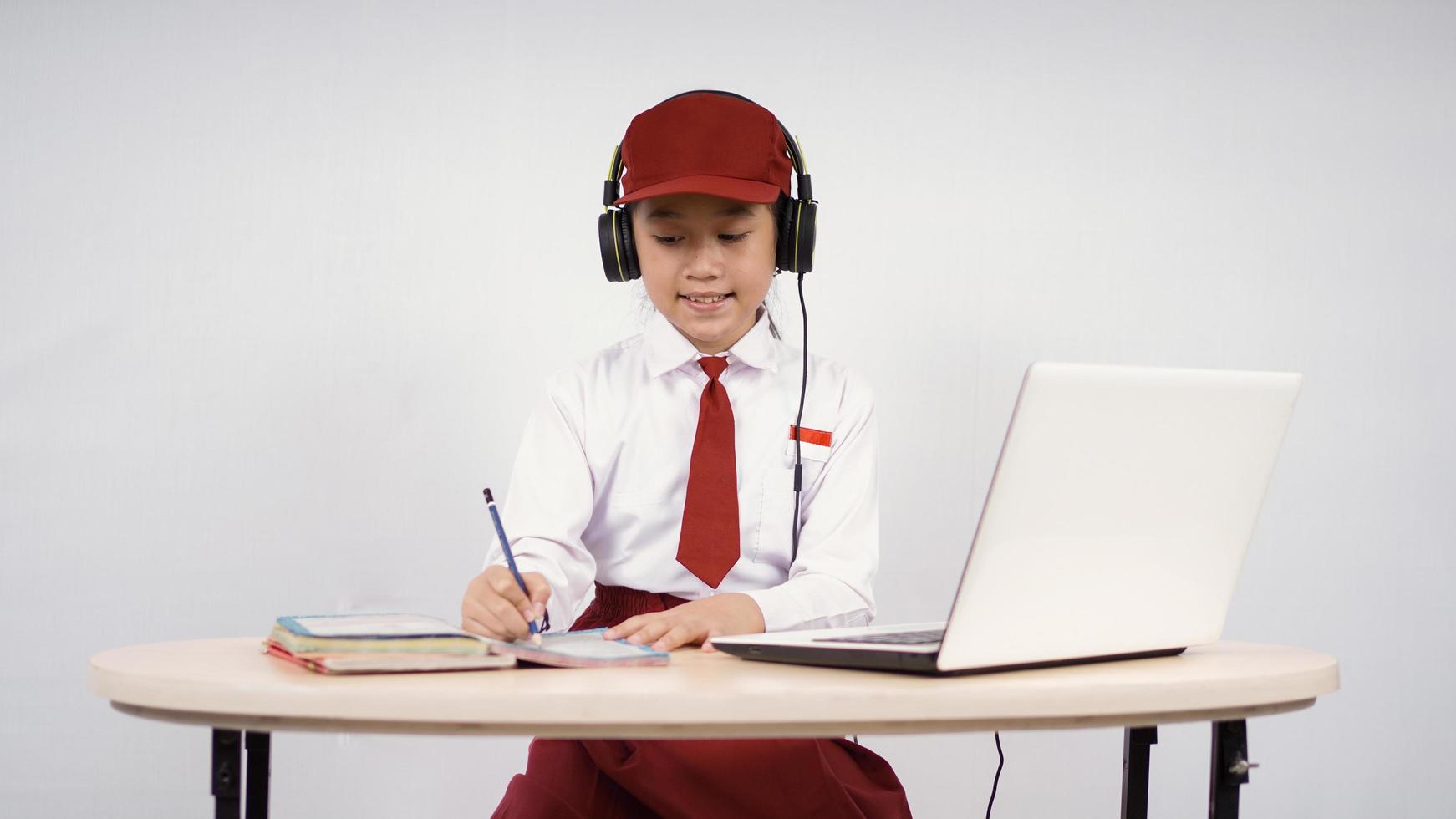 Elementary school asian girl listening on headphones while writing isolated on white background photo