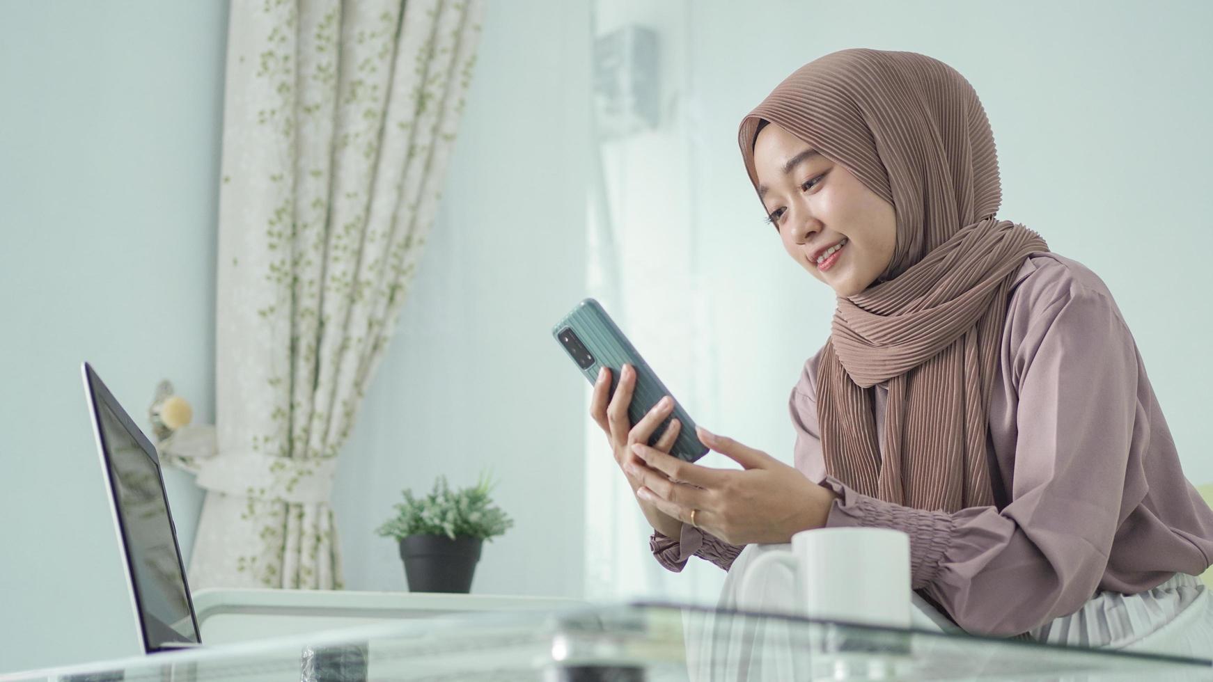 asian woman in hijab looking at phone screen happily at home photo