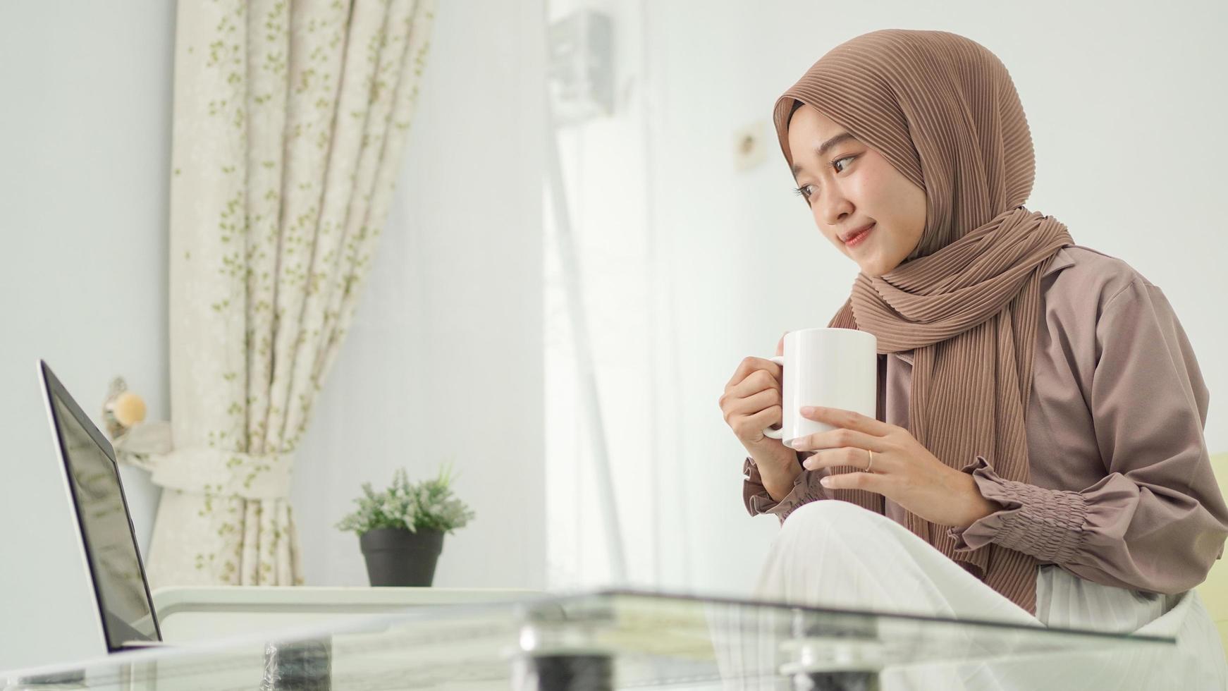 asian woman in hijab working from home using laptop while enjoying a drink photo
