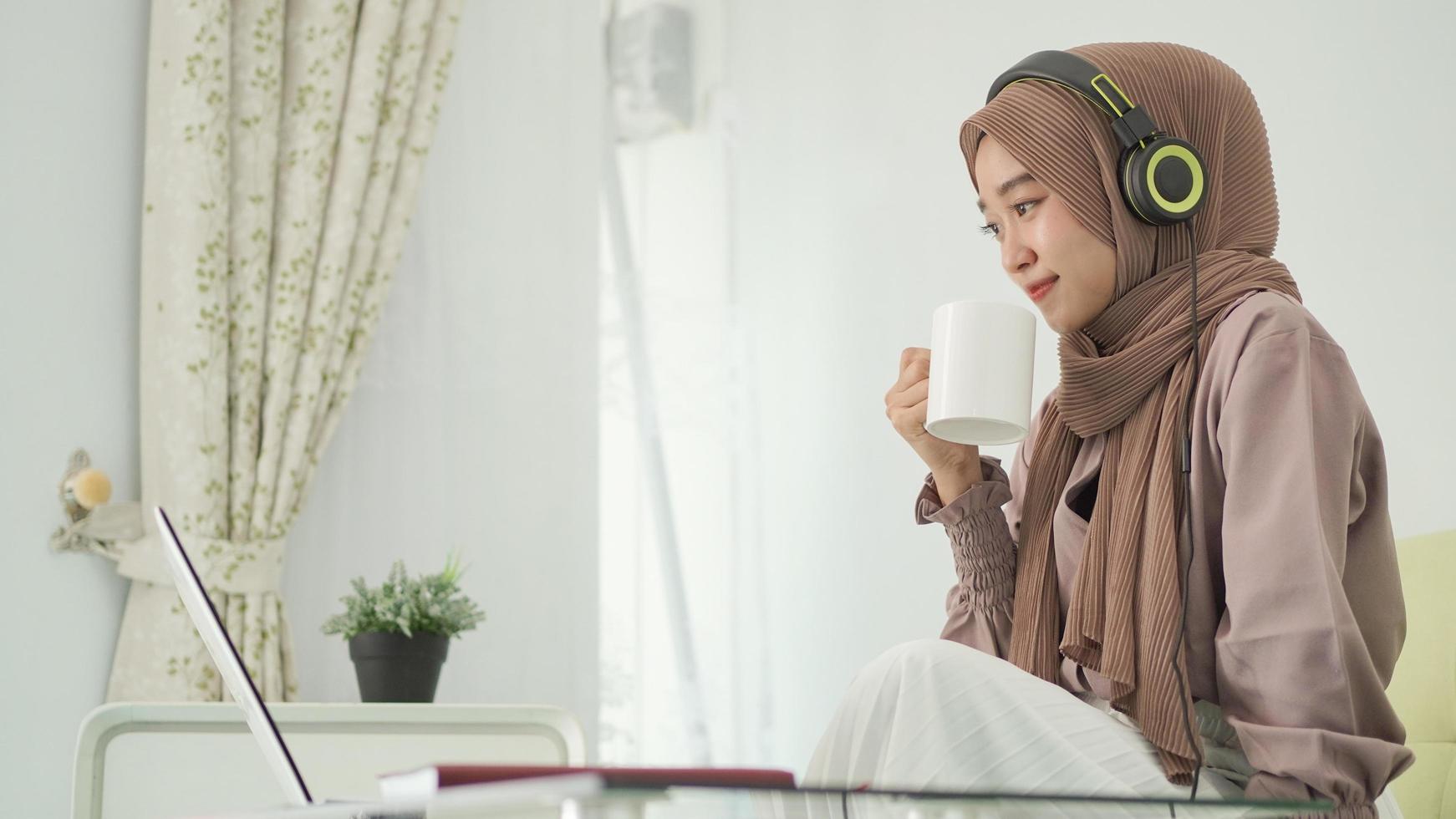 asian woman in hijab working from home enjoying a drink while listening photo