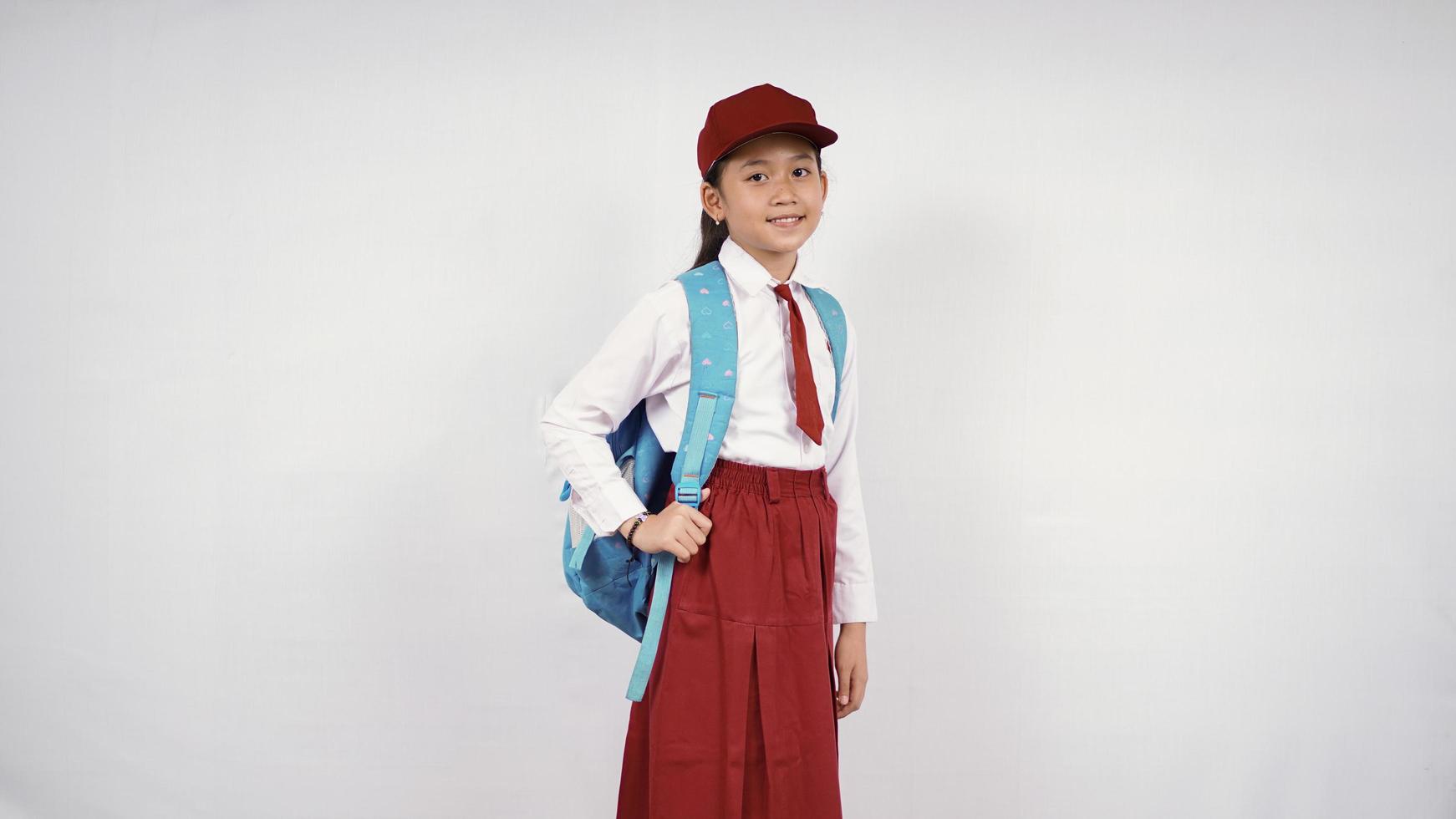 niña asiática con sombrero y mochila escolar sonriendo alegremente con fondo blanco aislado foto