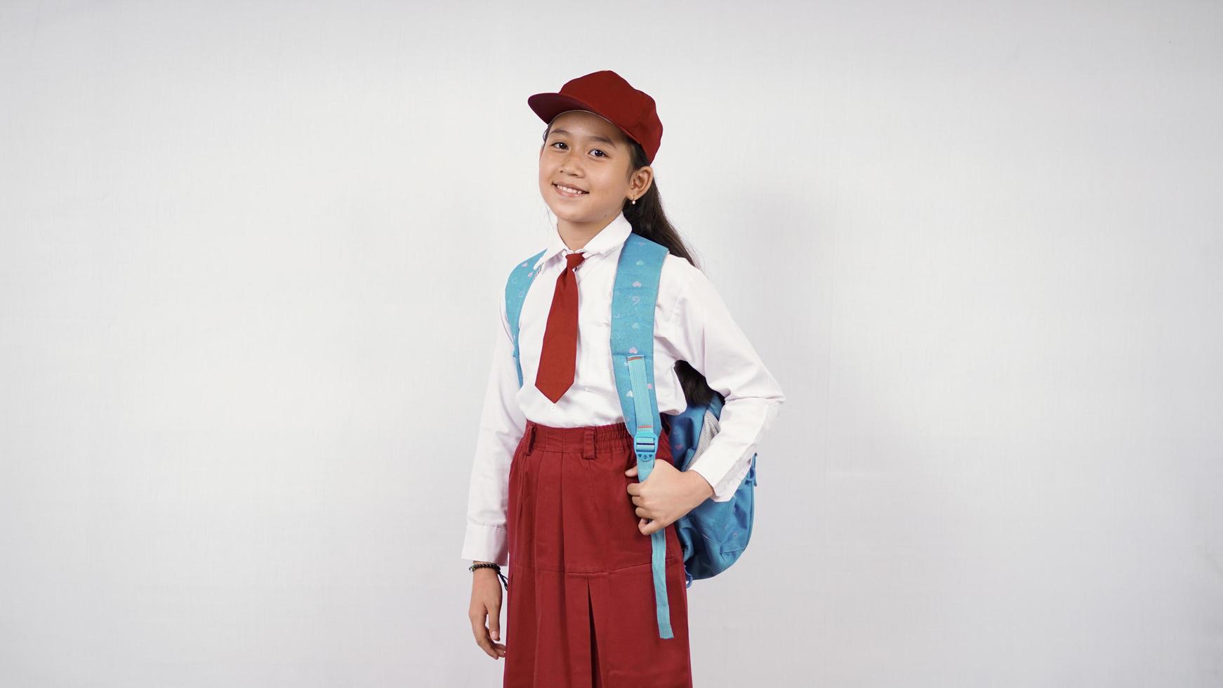 niña asiática con sombrero y mochila escolar sonriendo alegremente con fondo blanco aislado foto