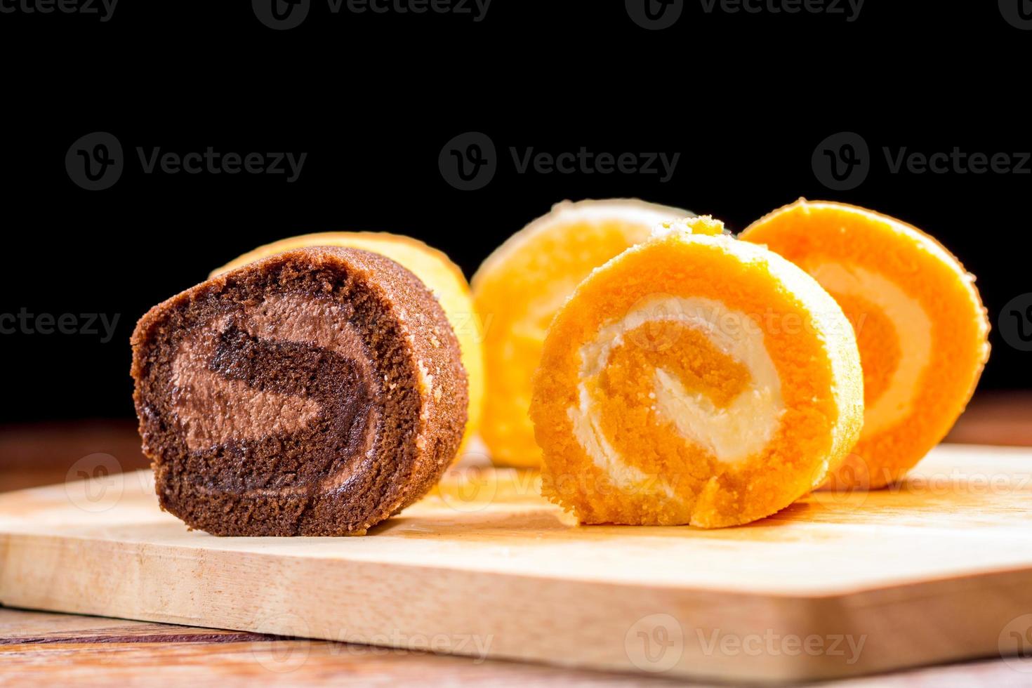 chocolate, vanilla and orange roll cake with whipping cream on  a wooden chopping board and table at the home kitchen with a black background and copy space. Homemade bakery concept. photo