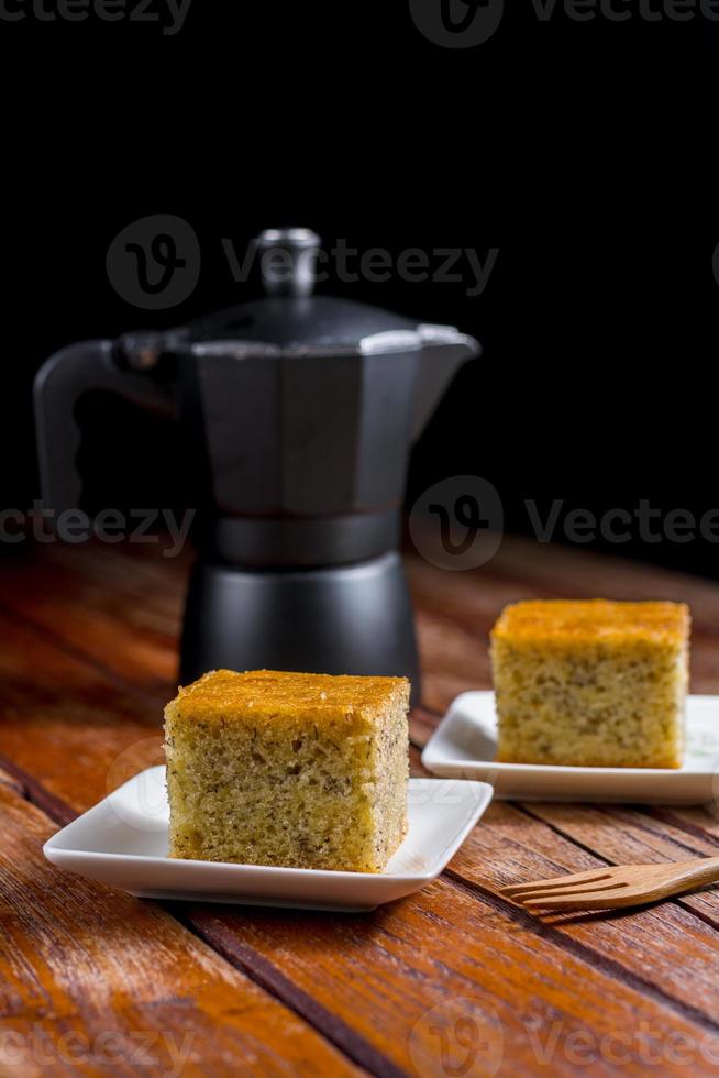 Close up square cut of homemade sweet and solf banana cake on white plate on table with solf focus black moka pot. Delicious and healthy bakery. photo