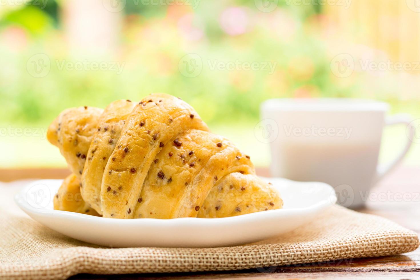 concepto de desayuno. croissants con semillas de perilla en un plato blanco y una taza blanca de café negro en una mesa de madera con fondo verde de bokeh. foto