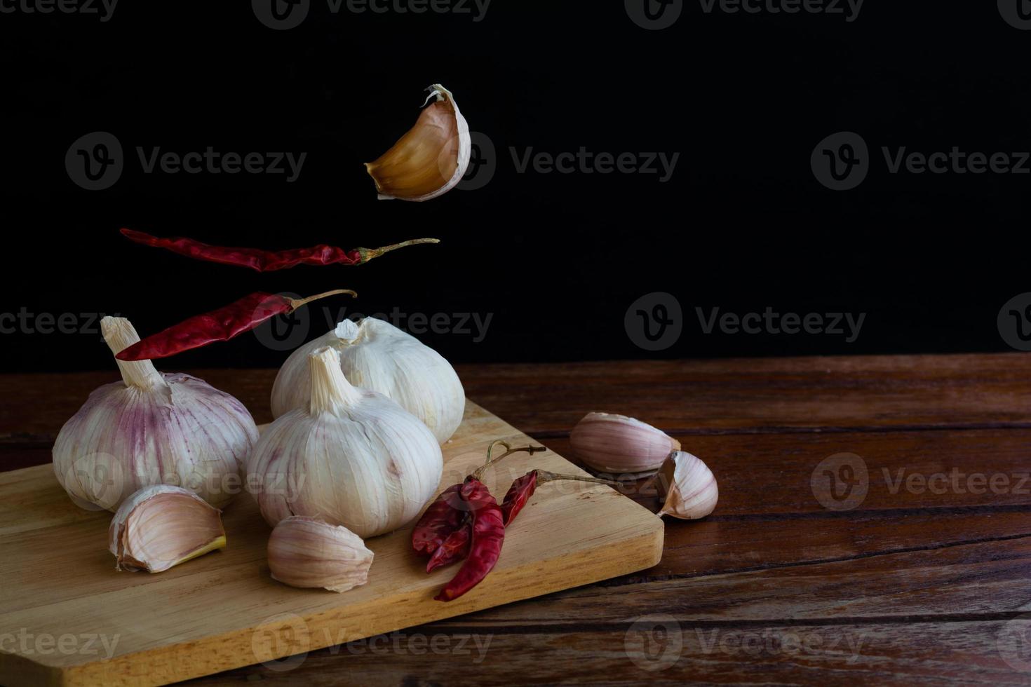 Group of garlic on chopping board and some garlic cloves floating in the air and red dried chilli on wooden table with black background. Copy space for your text. photo